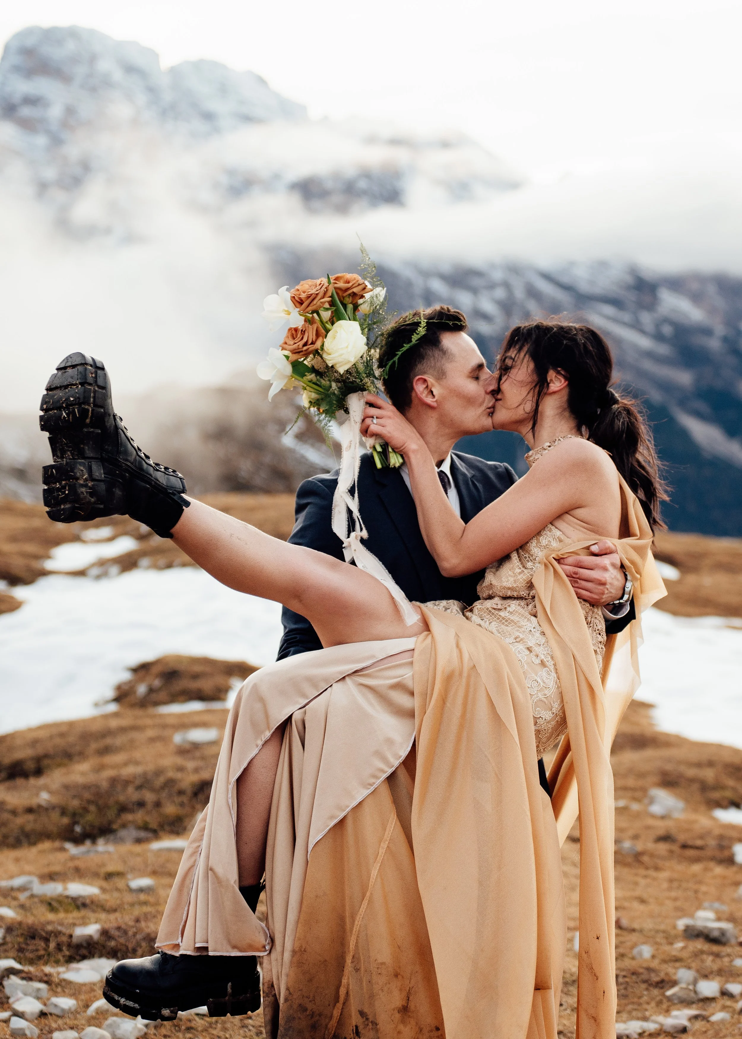 Couple eloping at tre cime di lavaredo sharing kiss wearing wedding dress holding bouquet of flowers while husband holds wife in arms