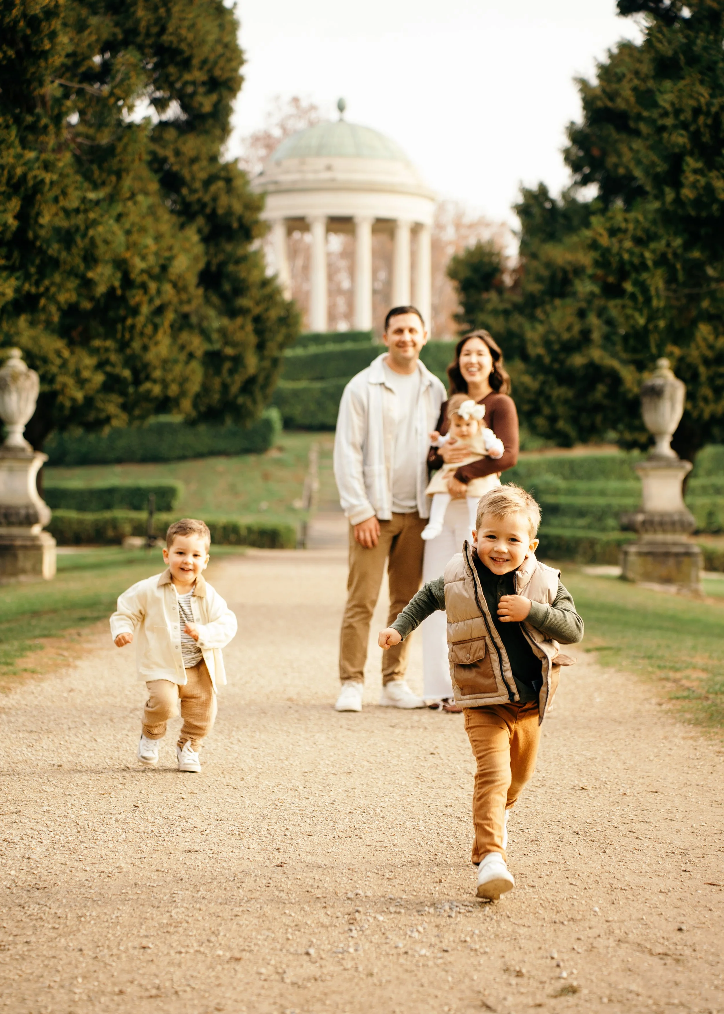 young brothers running playfully toward the camera on a path with parents in a classic Italian villa garden.