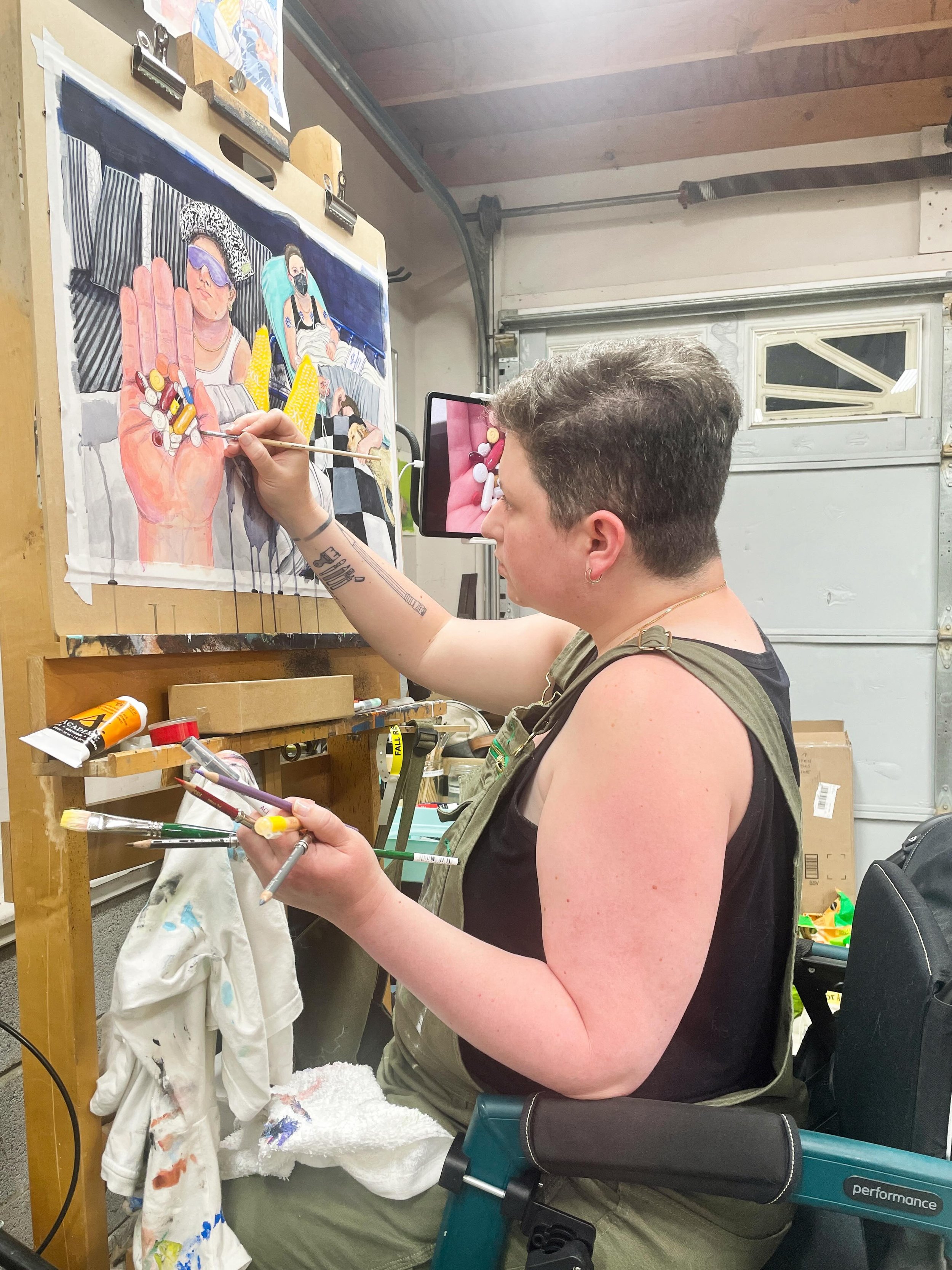 Just a little work in progress studio photo. 
&nbsp;
accessibility: A photo of Ashley seated in her teal Rollz Motion Performance rollator, wearing a green pair of overalls and a black tank top with, holding up a paintbrush in her right hand to a pai