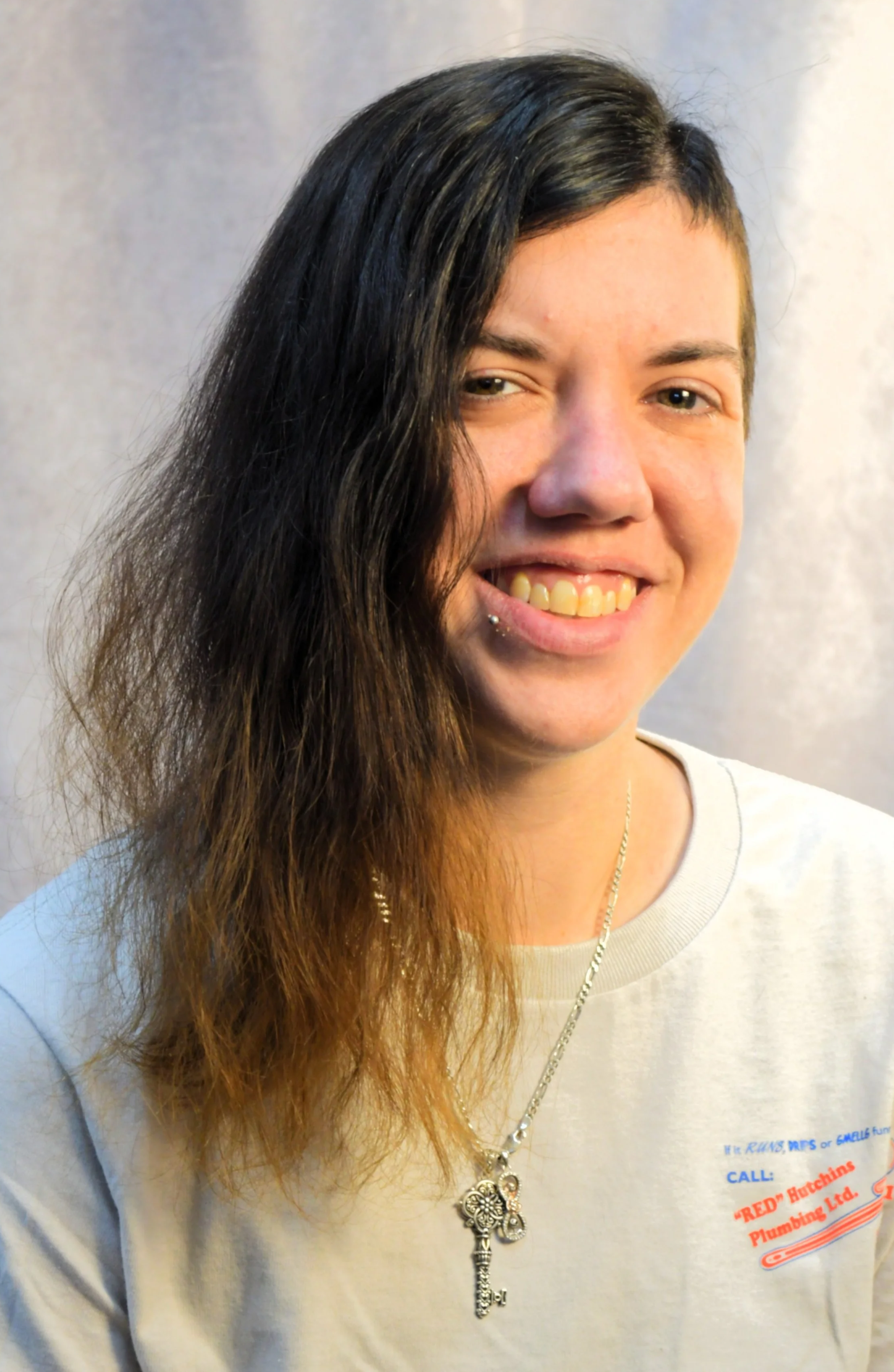 A woman with black and brown wavy hair, partially shaved on the right side, wearing a light-colored t-shirt with red and blue text, a silver necklace with a key pendant, and a lip piercing, smiling at the camera.