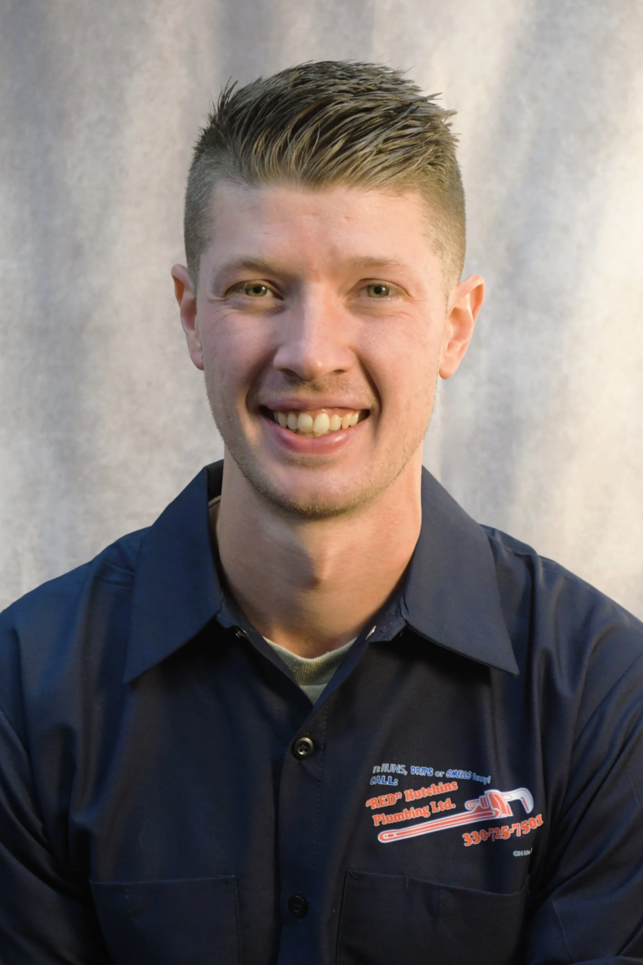 Young man smiling, wearing a dark blue shirt with a company logo for plumbing services.