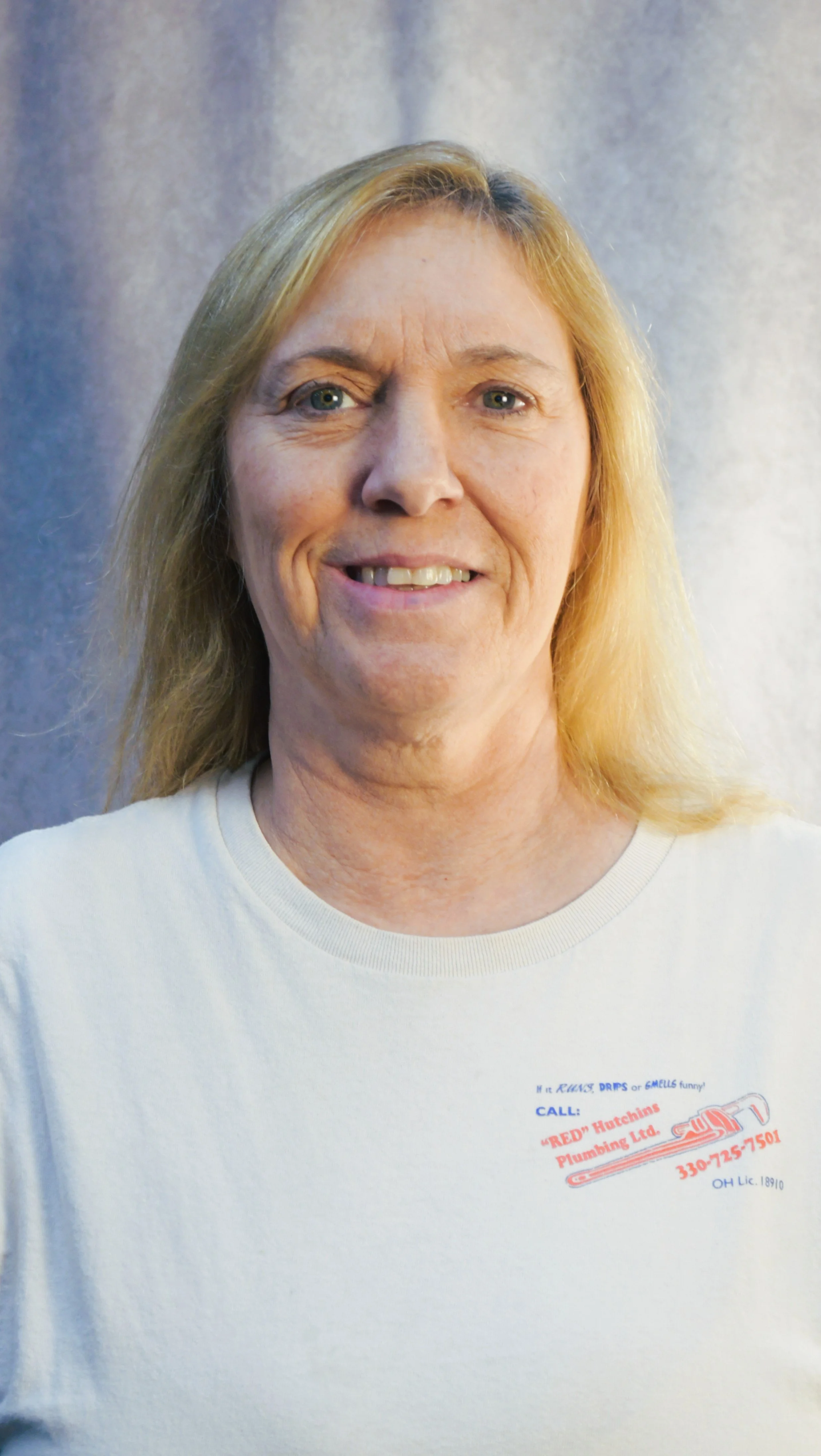A woman with shoulder-length blonde hair smiling, wearing a white T-shirt with a plumbing company logo and contact information.