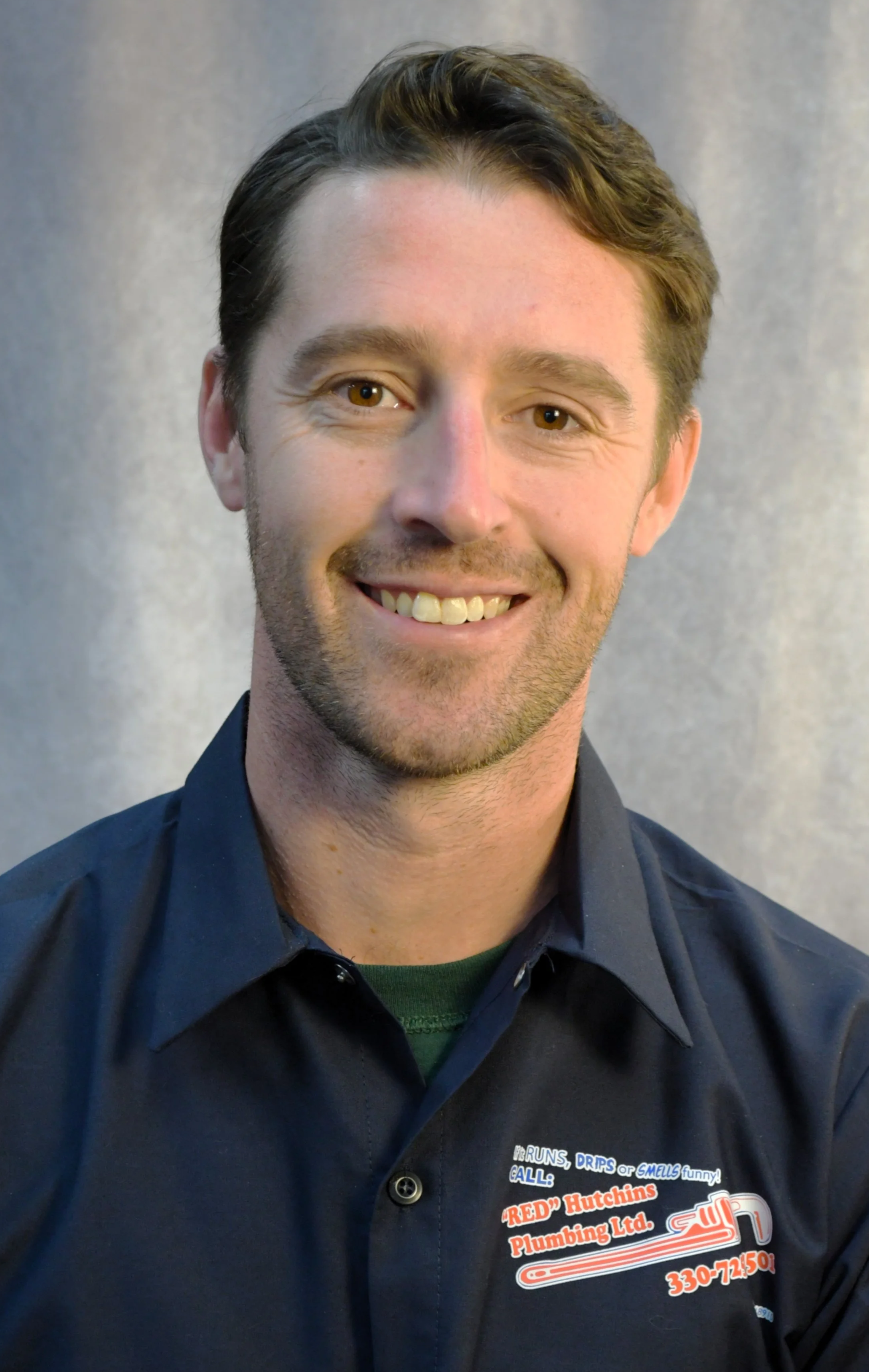 Portrait of a smiling man with brown hair, wearing a navy blue button-up shirt with a plumbing company logo.