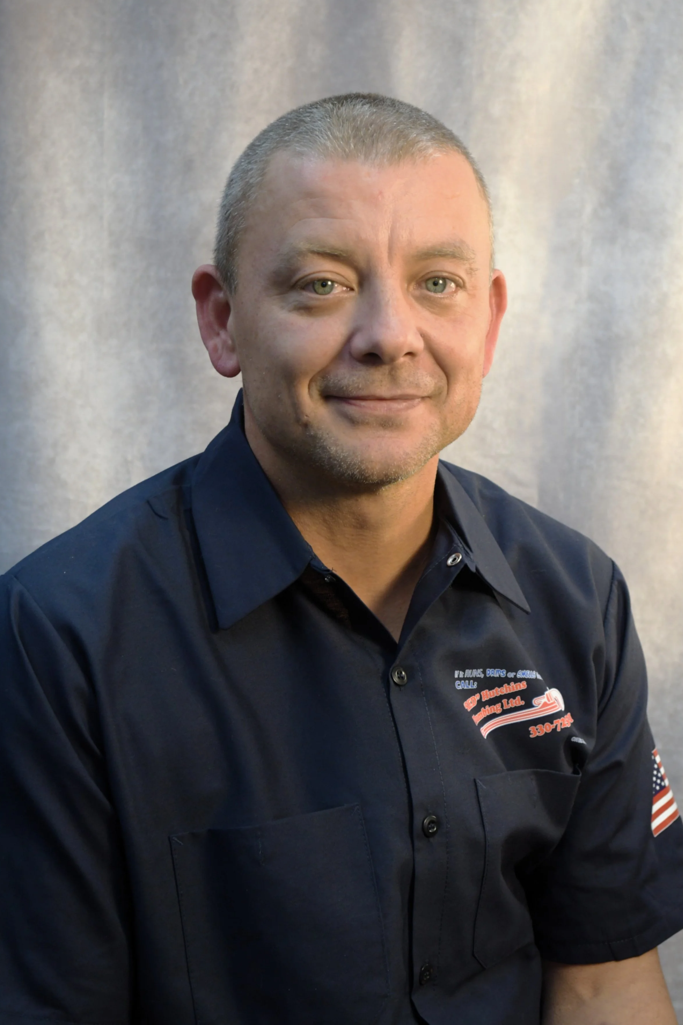 A man with short hair wearing a dark blue work shirt with company branding, smiling, sitting against a light-colored background.