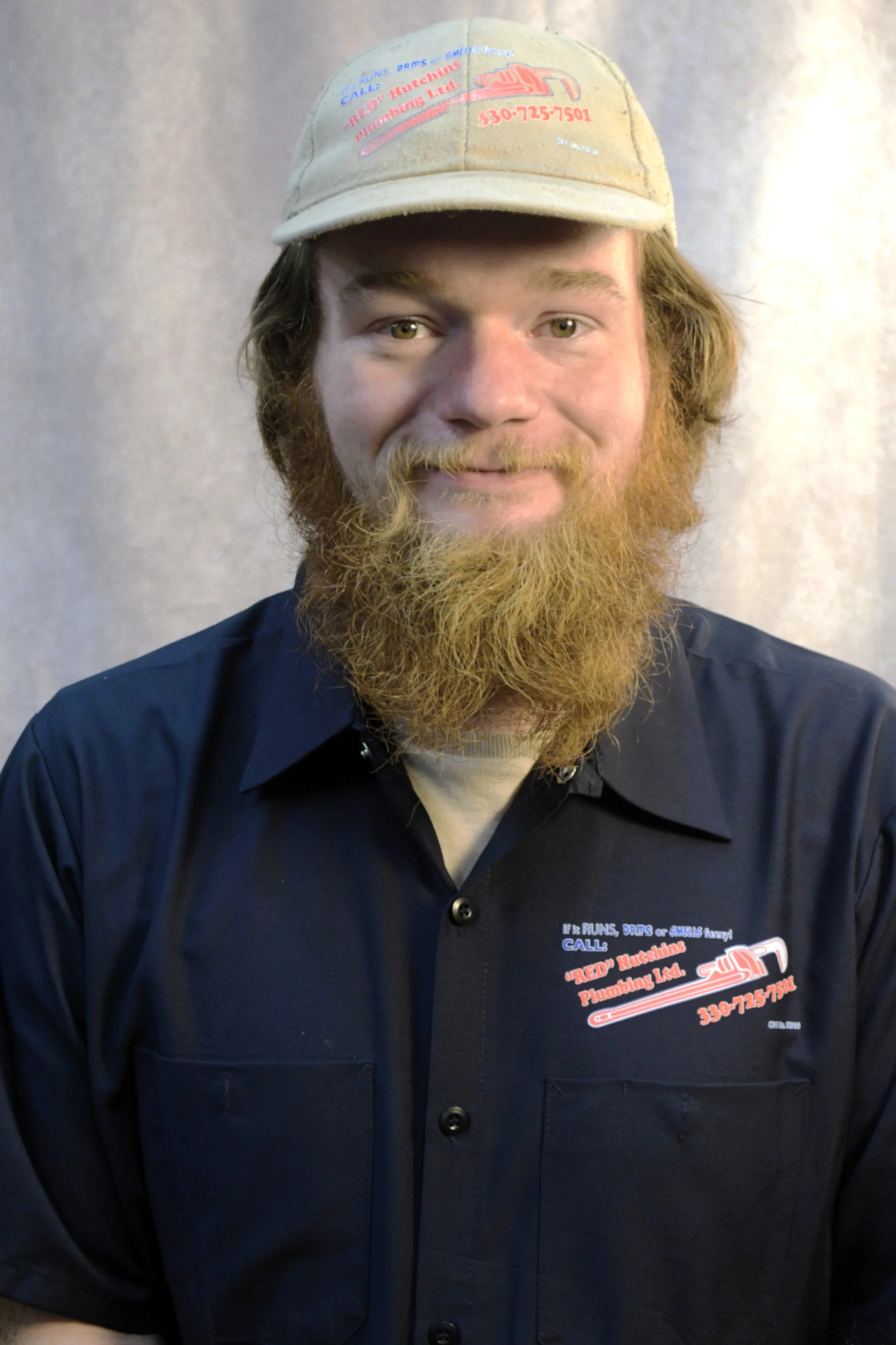 A man with a beard and mustache wearing a cap and a navy blue shirt with the logo 'Red Hutchins Plumbing Ltd.' on it, standing in front of a plain background.