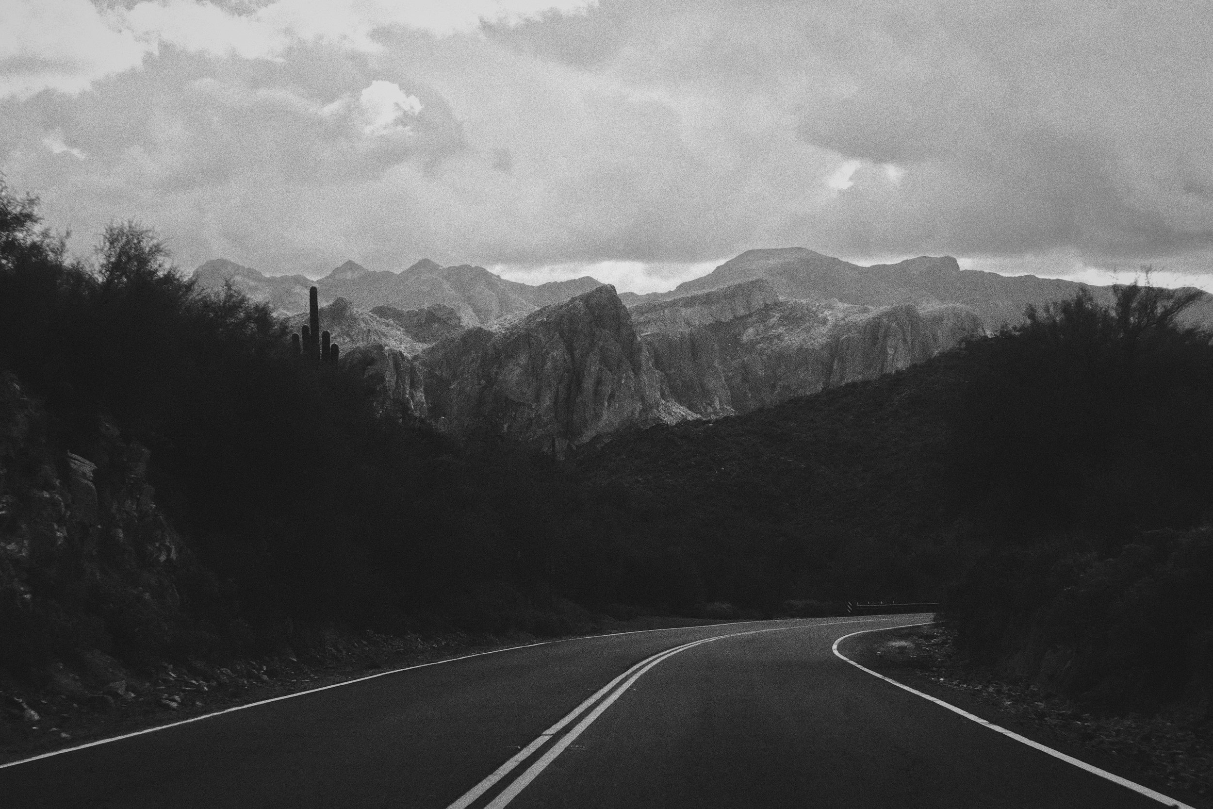 A winding road through a mountainous desert landscape with cacti, rocks, and trees, under a cloudy sky.