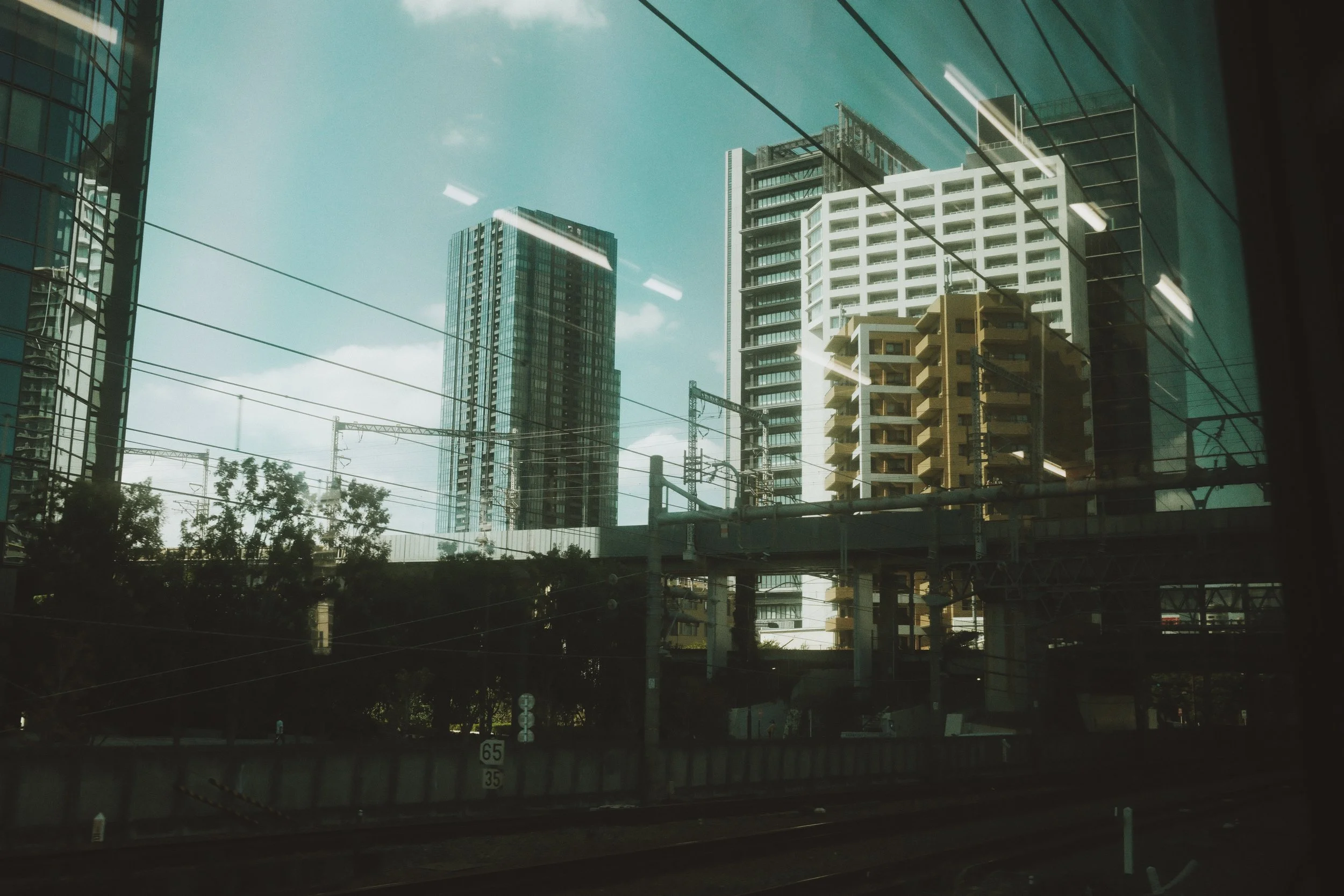 View of tall modern skyscrapers and high-rise buildings through a glass window, with reflections of lights and electrical lines in the foreground.