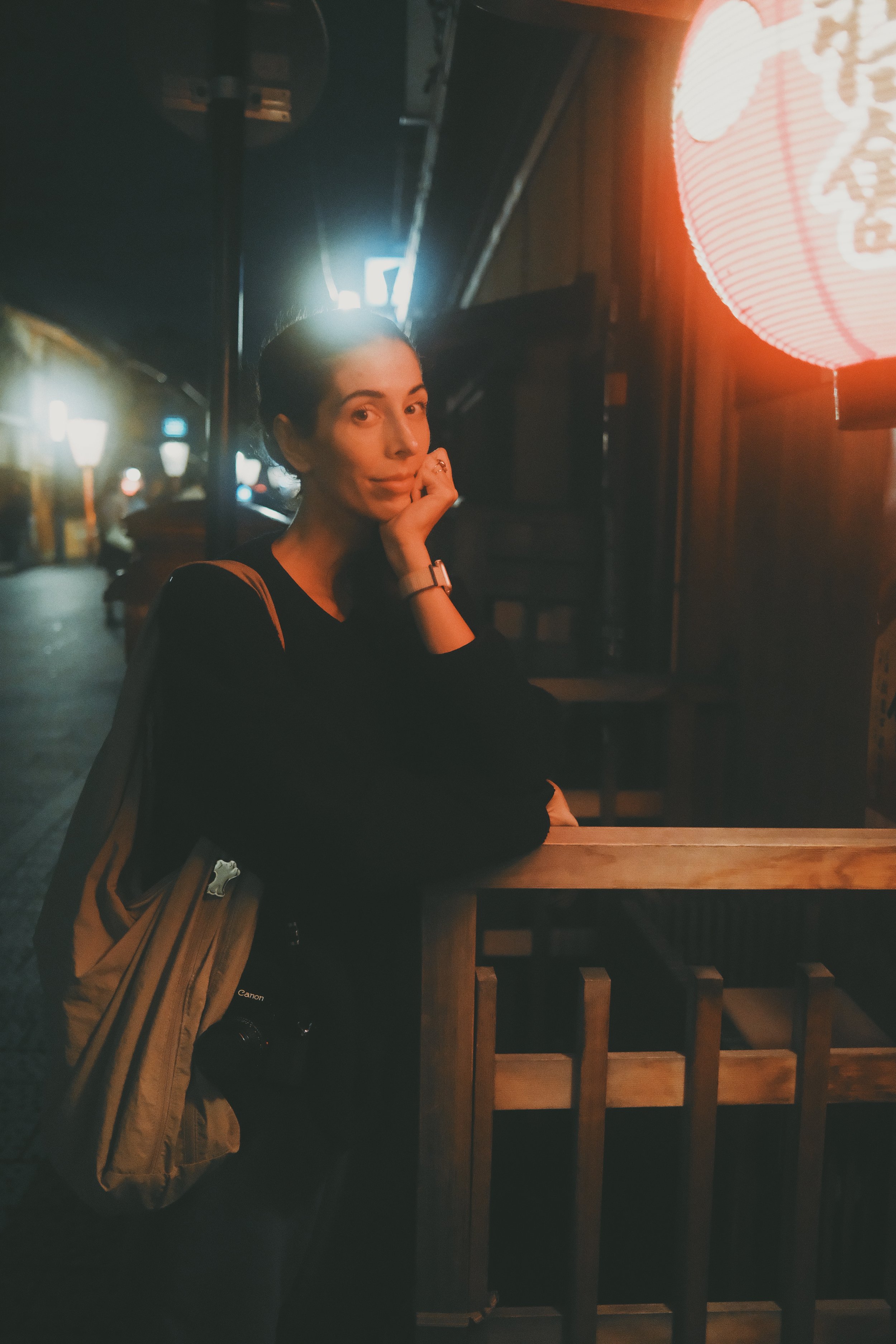 A woman standing inside a dimly lit restaurant or bar, resting her chin on her hand, with a camera hanging from her shoulder, illuminated by warm, red-orange lantern light.