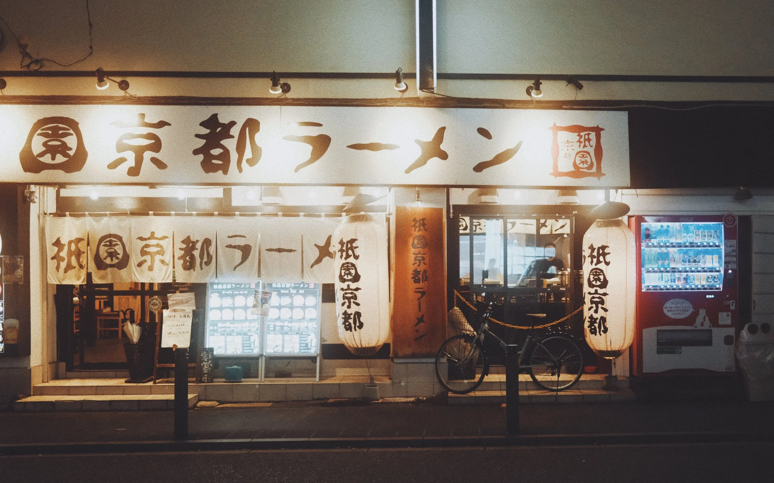 Front of a Japanese ramen restaurant at night with traditional lanterns, a bicycle parked outside, and vending machines to the right.