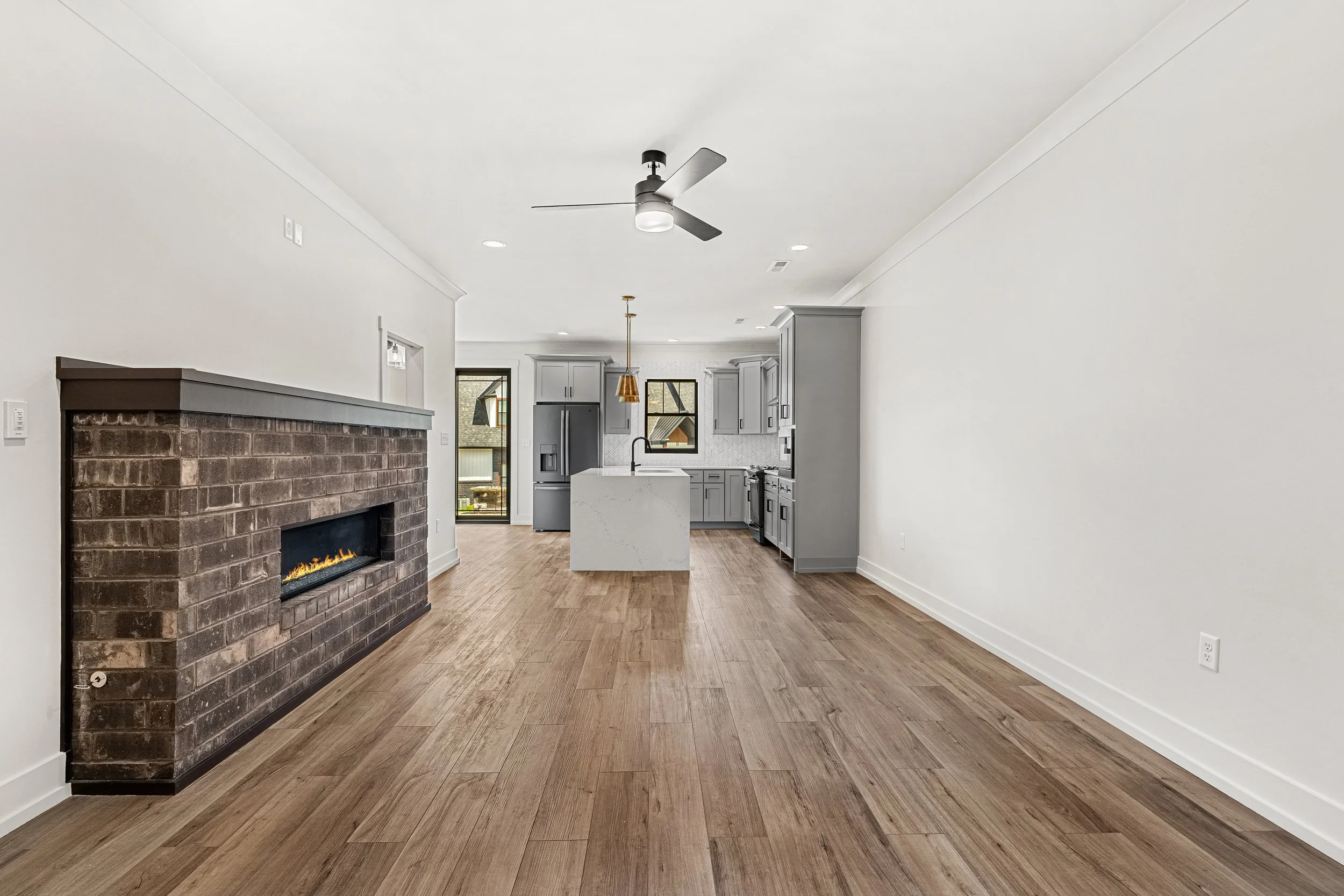 Modern kitchen with light gray cabinets, white countertop island, black hardware, black stainless steel appliances, wood floor, and a window showing neighboring houses.