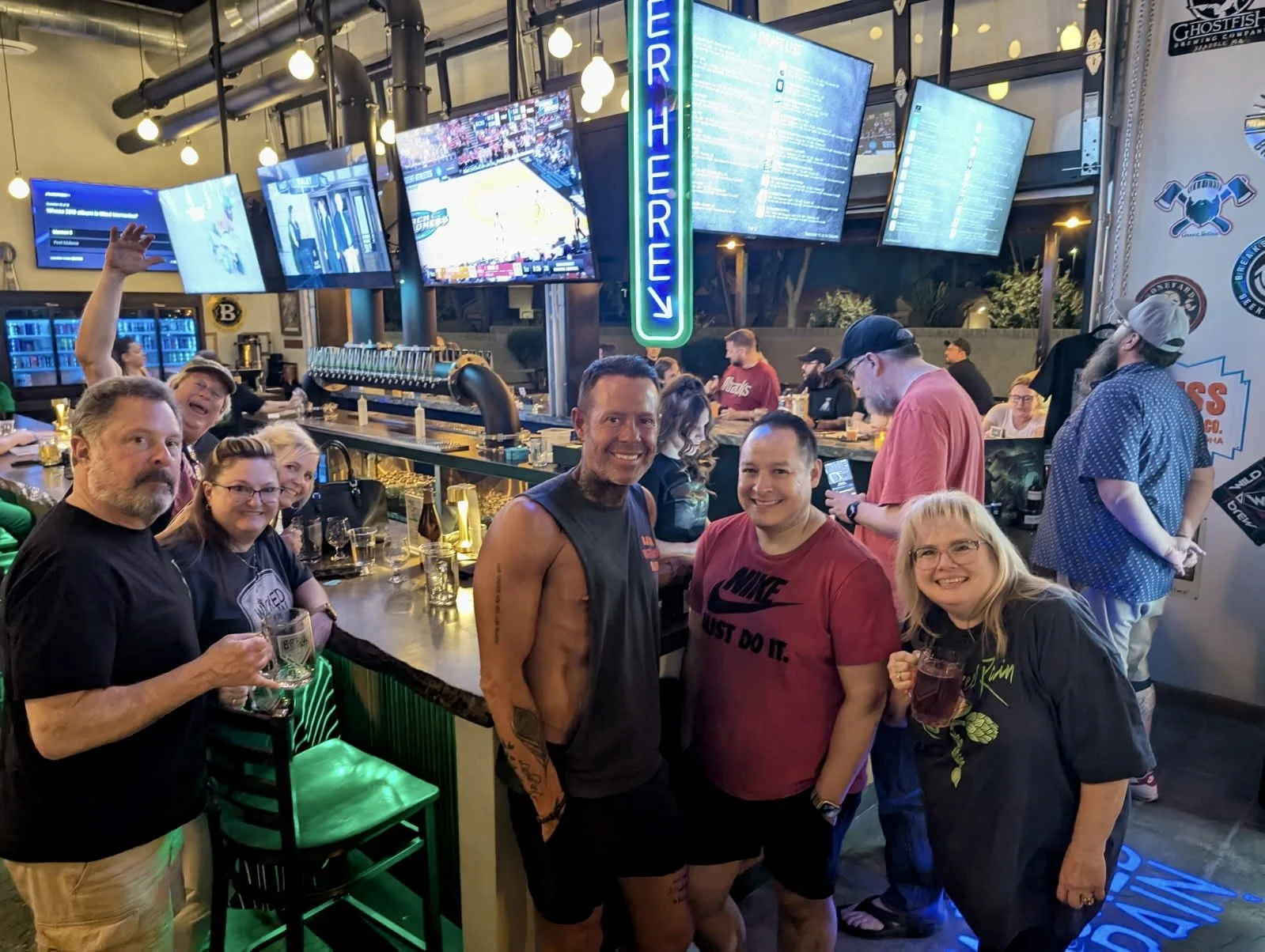 A group of people at a bar celebrating, some holding drinks, with multiple TV screens at Wicked Rain