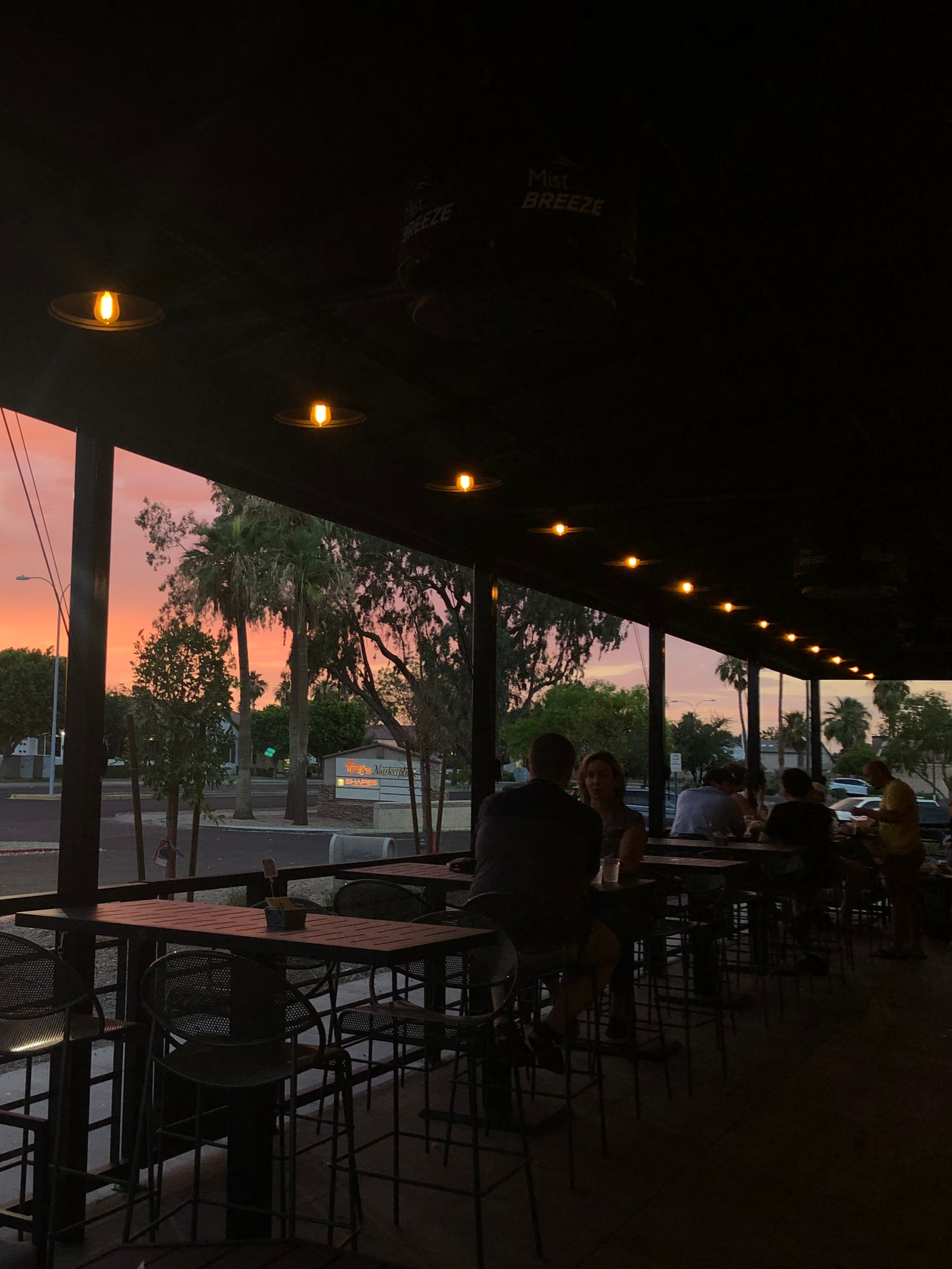 People sitting at tables on the patio of Wicked Rain during sunset, with a view of trees and a parking lot outside.