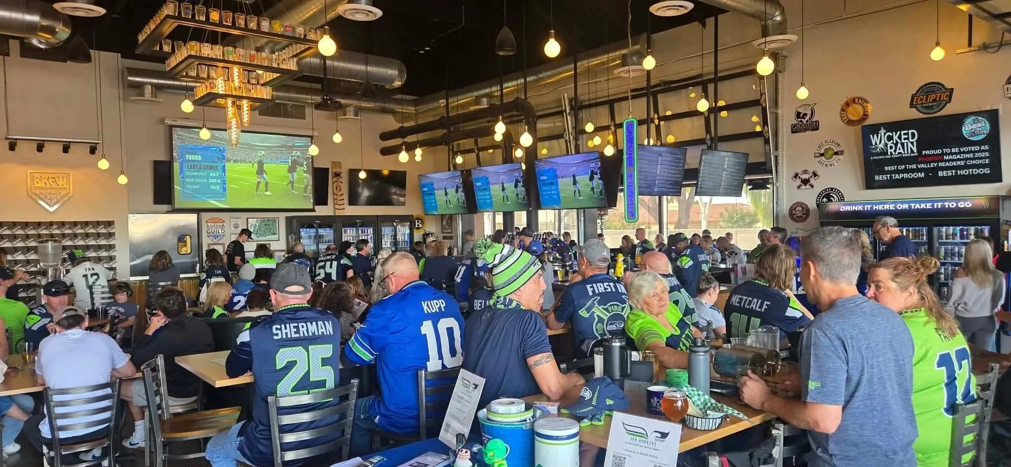 Inside of a Wicked Rain is filled with many people watching a football game on large screens. The crowd is wearing Seahawks jerseys and hats, eating and socializing. Multiple screens display the game, and tin tackers are displayed on the wall.