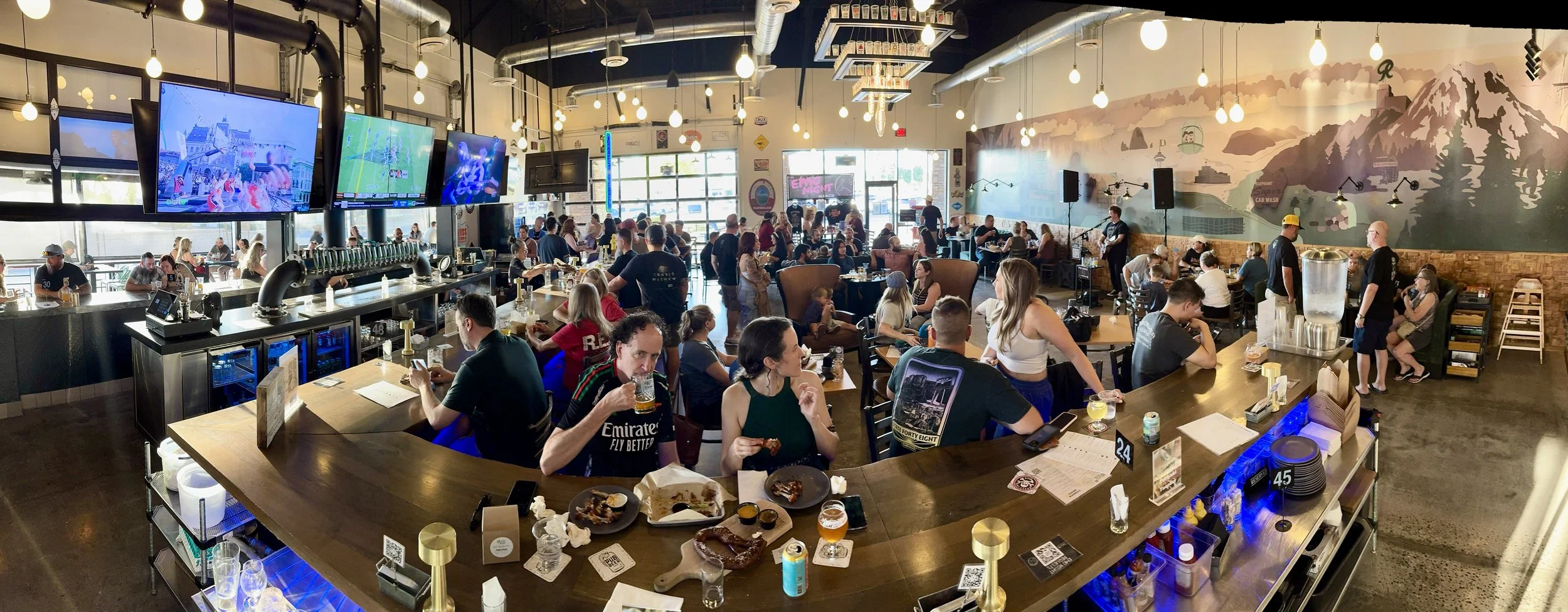 Crowded bar with patrons watching multiple large TV screens, some drinking, eating, and socializing during live music night at Wicked Rain