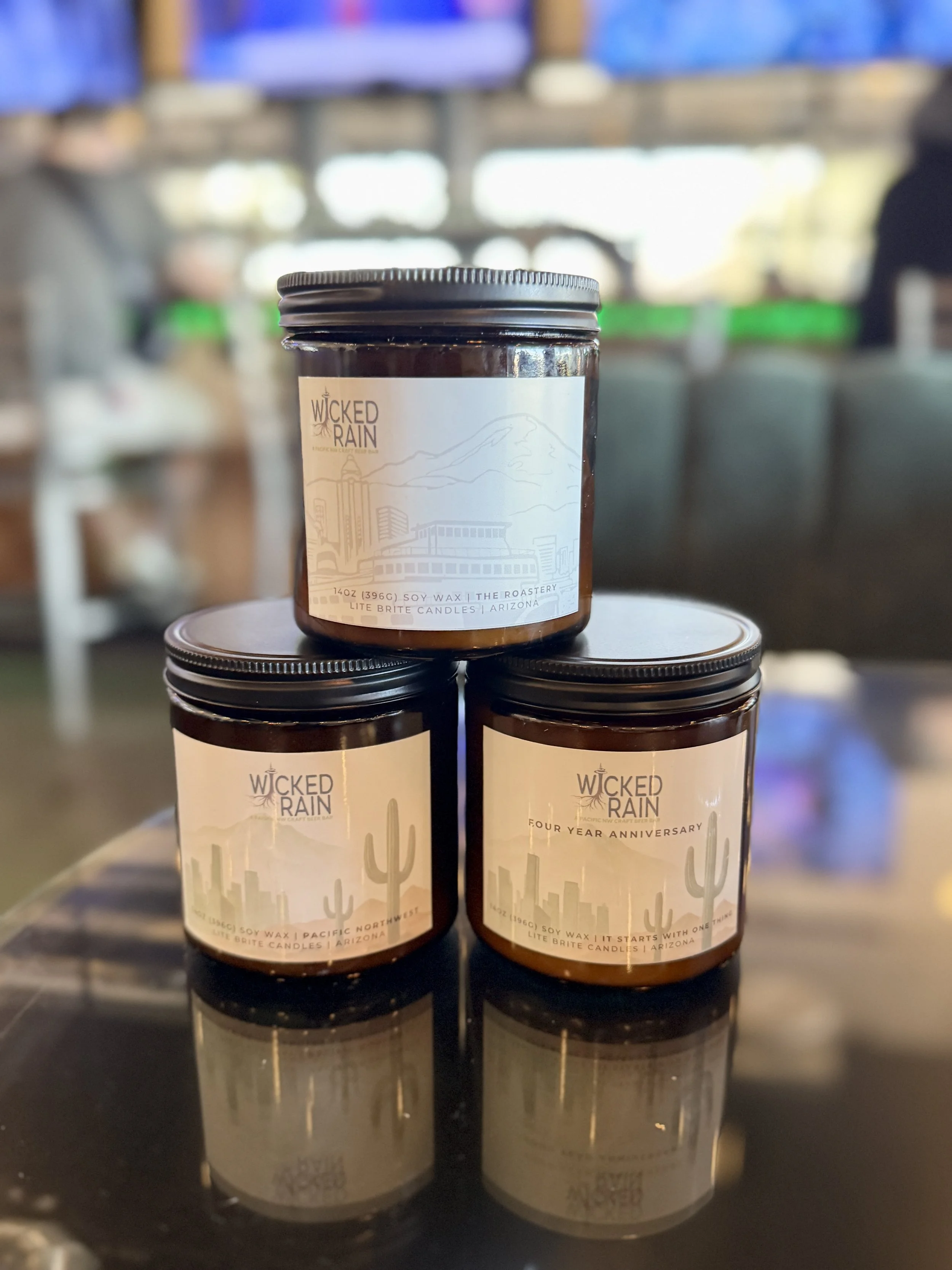 Three jars of Wicked Rain soy wax candles with desert-themed labels, placed on a reflective surface inside a cafe or similar setting.