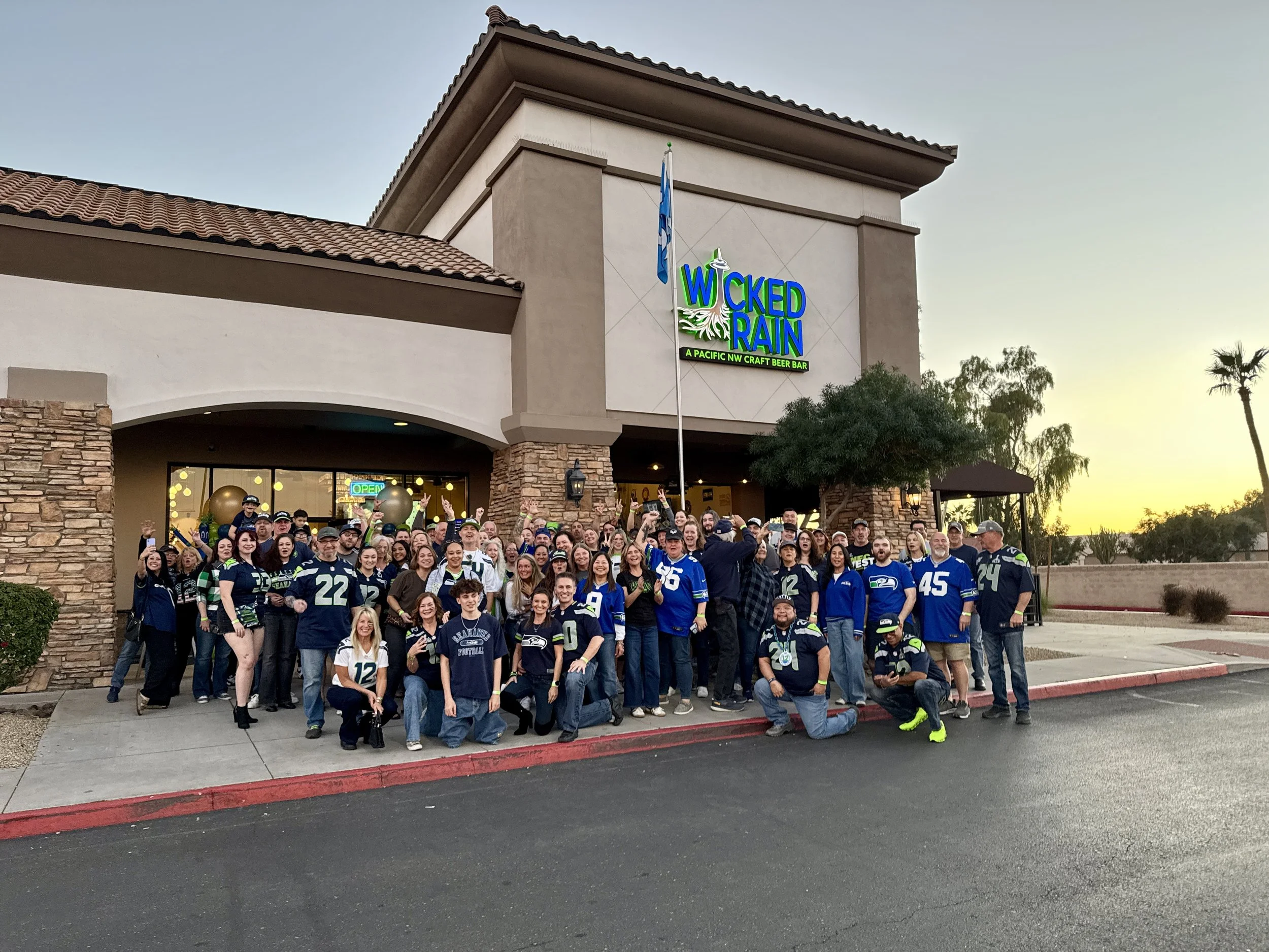 A group of people wearing Seattle Seahawks jerseys and attire gathered outside a restaurant called 'Wicked Rain' during sunset.