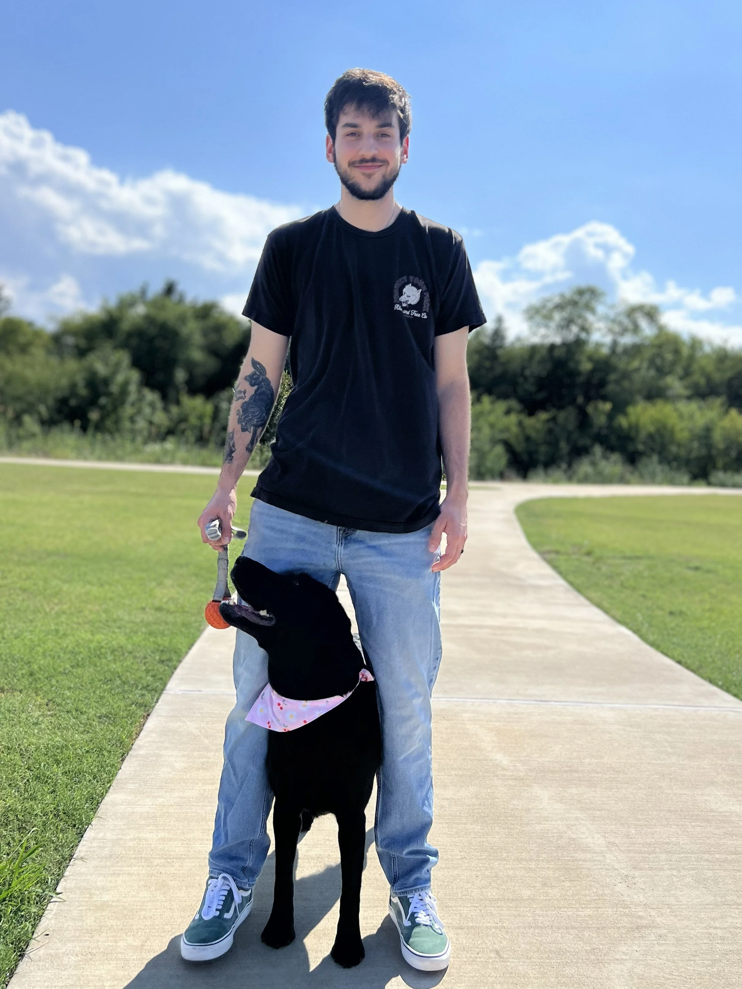 A young man with dark hair and beard standing on a sidewalk in a park, holding a leash attached to a black dog wearing a pink bandana, with green trees and a blue sky in the background.