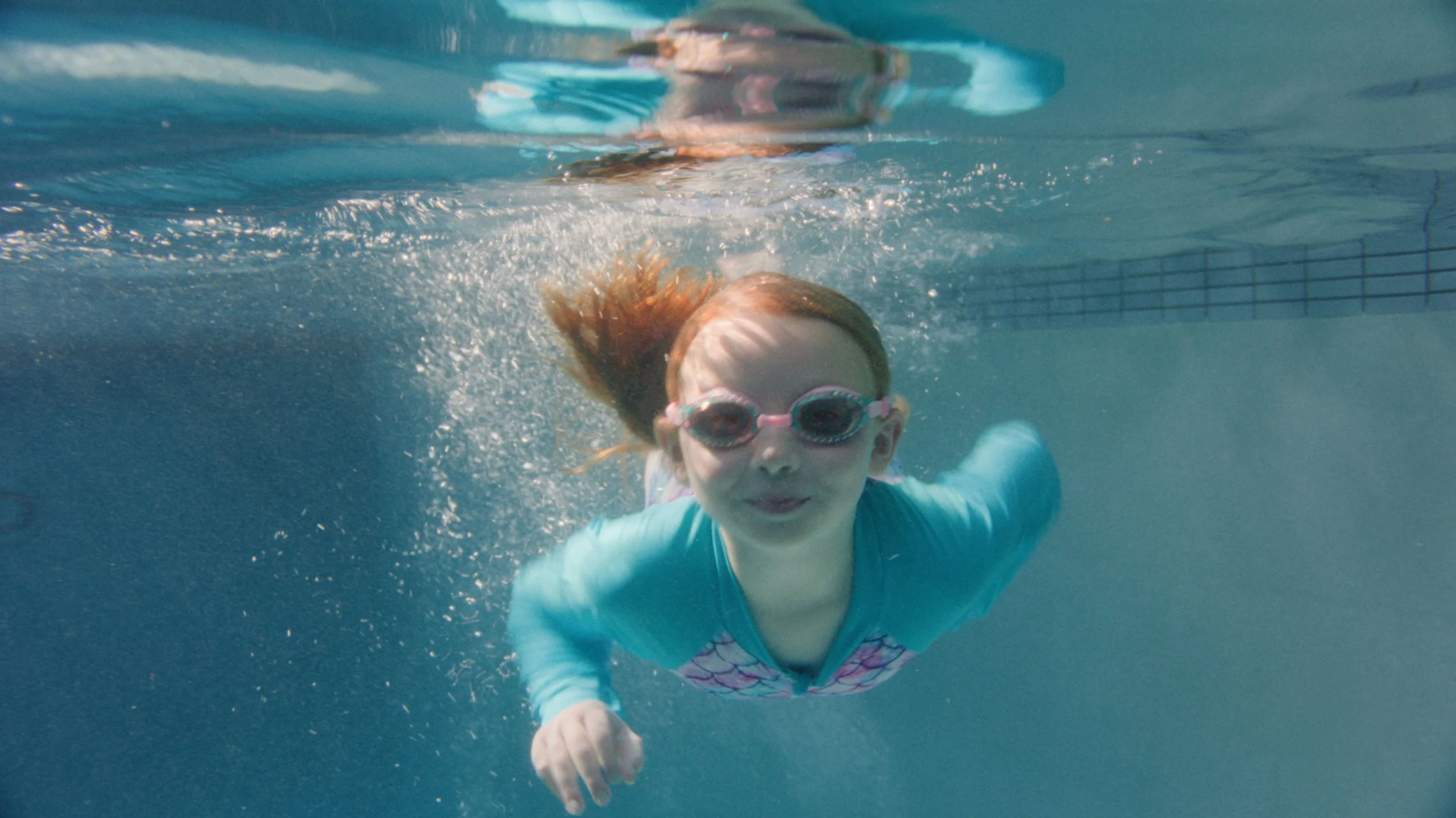 A young girl in swim goggles glides underwater through a sunlit pool, from Chase's All the Way Home films.