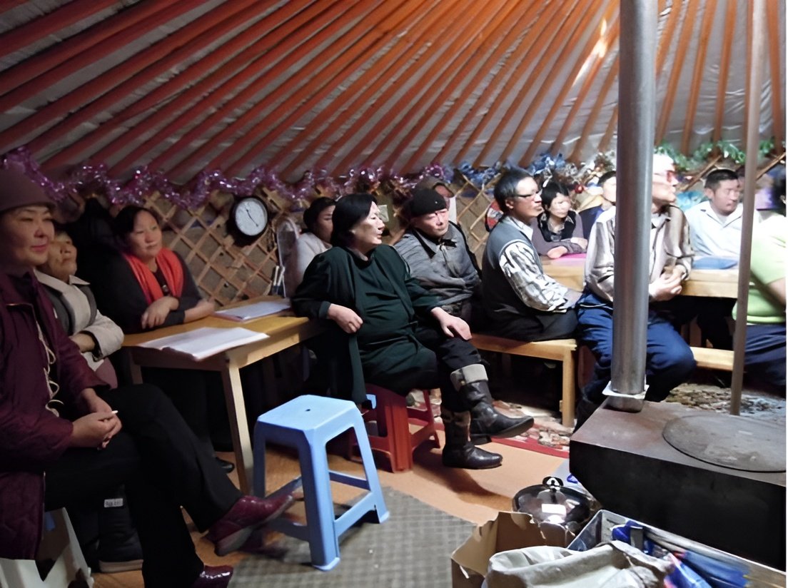 Group of people sitting inside a yurt, watching a performance or presentation, with traditional decorated interior walls.