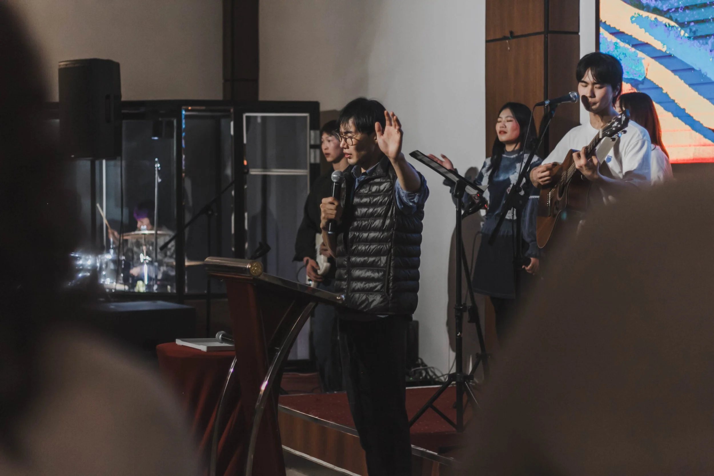 A group of young people performing music on stage, with one person singing into a microphone and others playing guitar and singing, in an indoor setting.