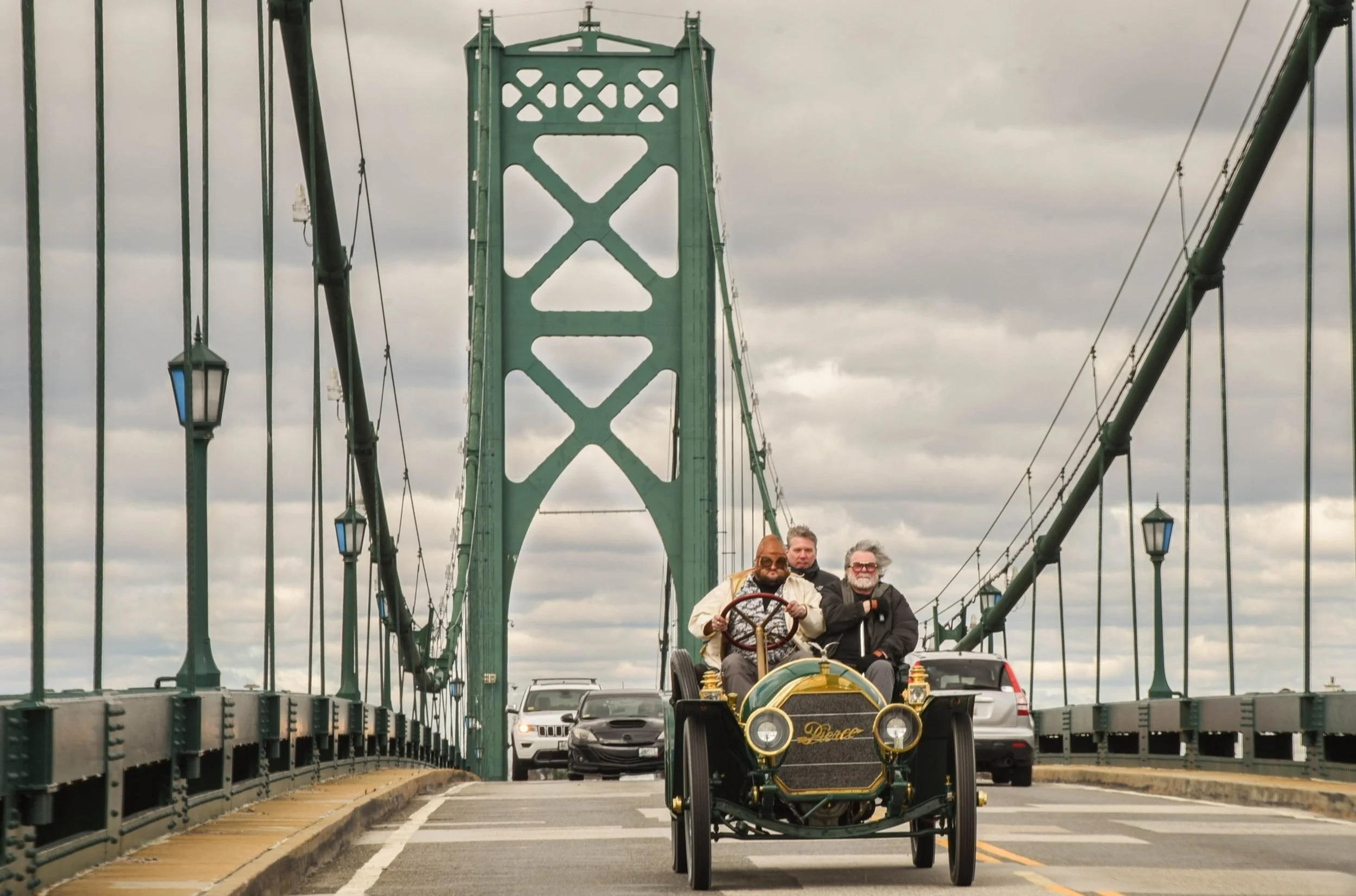 Three men riding in a vintage automobile across a bridge with green towers and suspension cables, cars following behind, cloudy sky above.