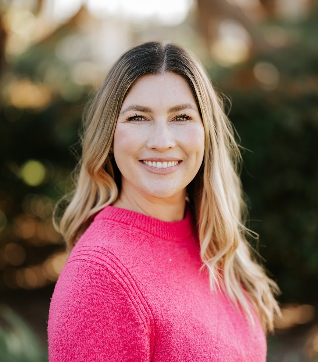 A woman with wavy blonde hair wearing a bright pink sweater, smiling outdoors with a blurred background of greenery and warm light.