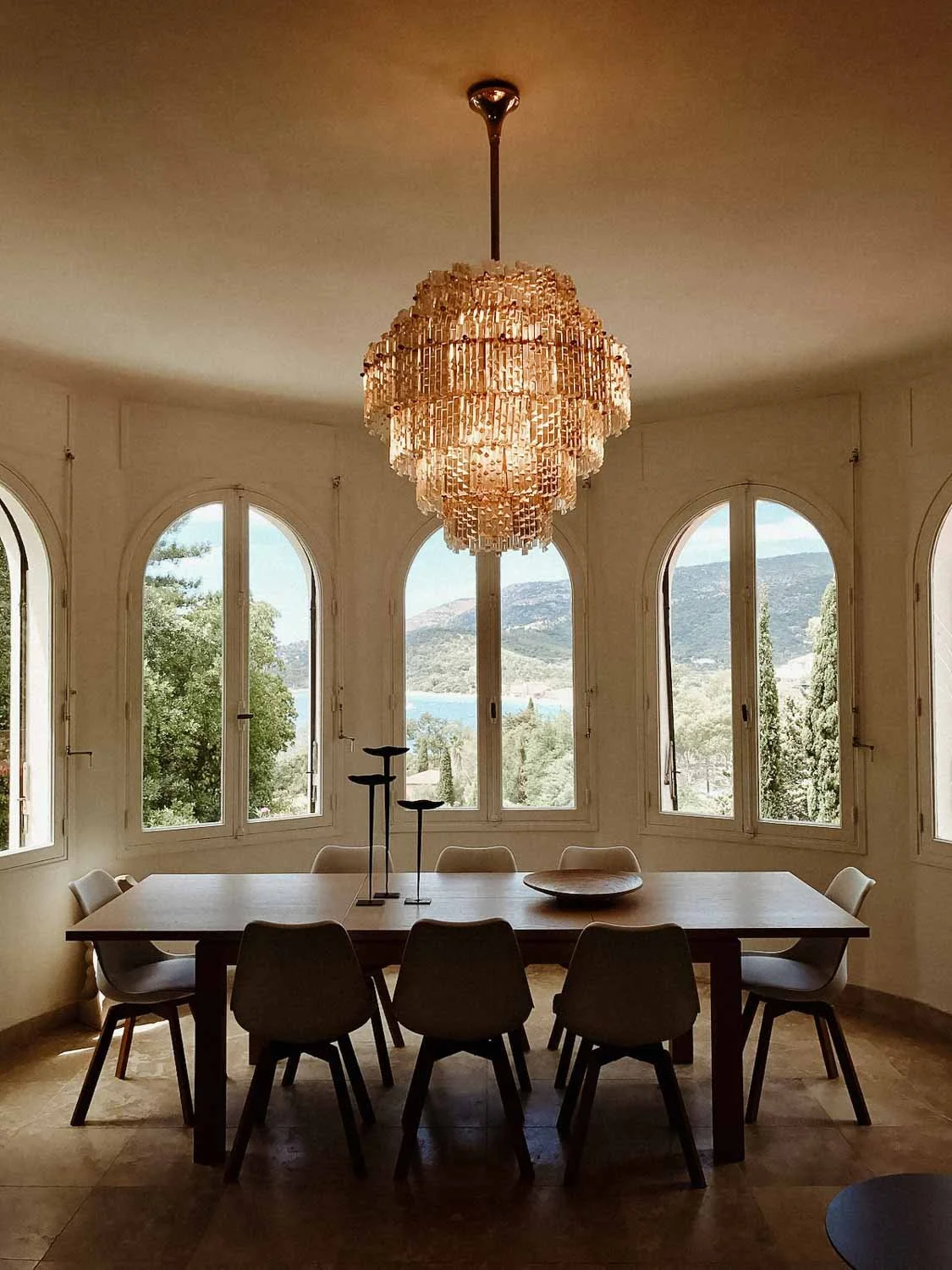 Salle à manger élégante avec grande table en bois entourée de chaises blanches, sous un lustre en cristal étincelant. Fenêtres en arc offrant une vue sur le paysage.