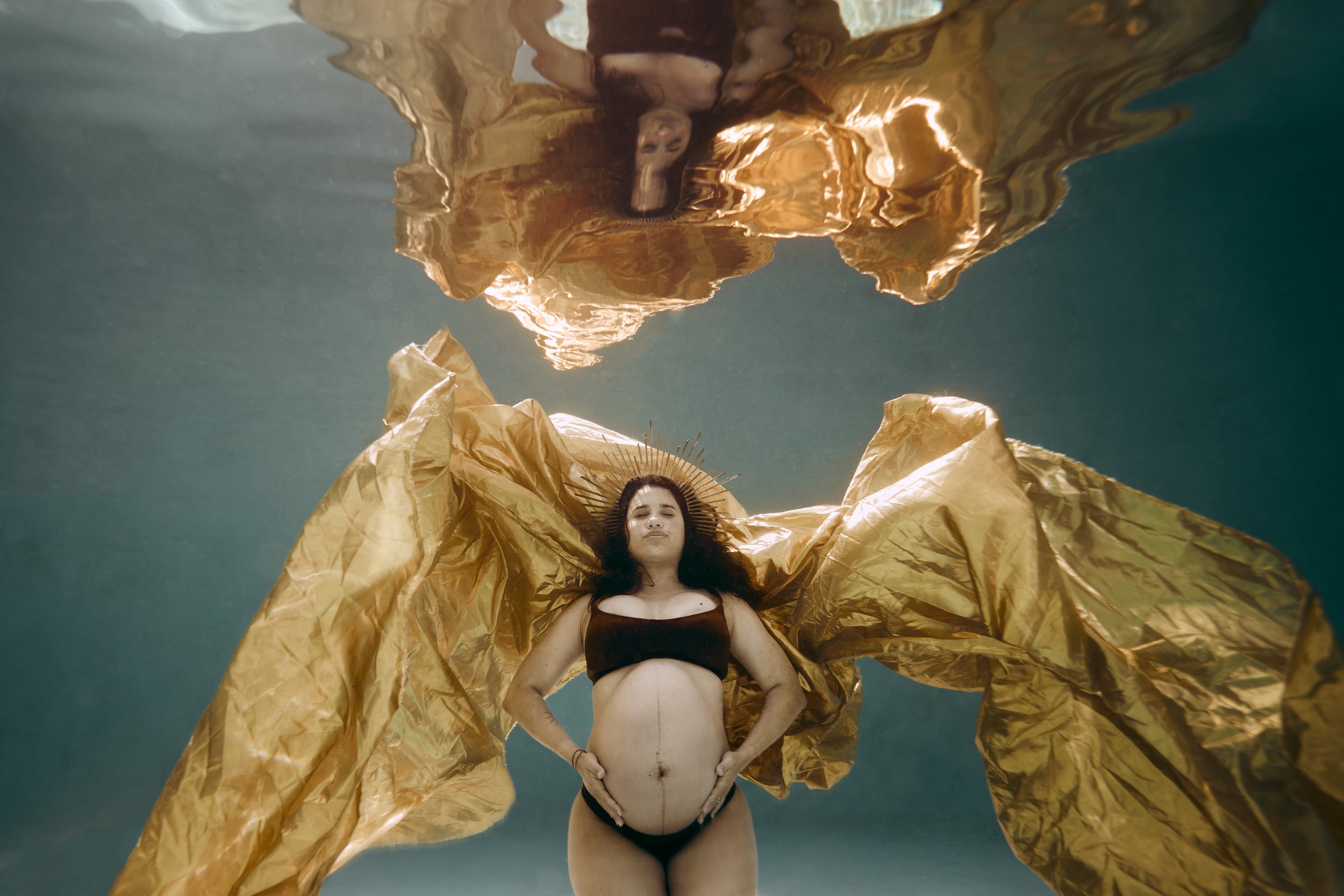 Future maman lors d'une séance photo aquatique. Elle est sous l'eau avec un drap doré. Telle une déesse de la beauté de la maternité