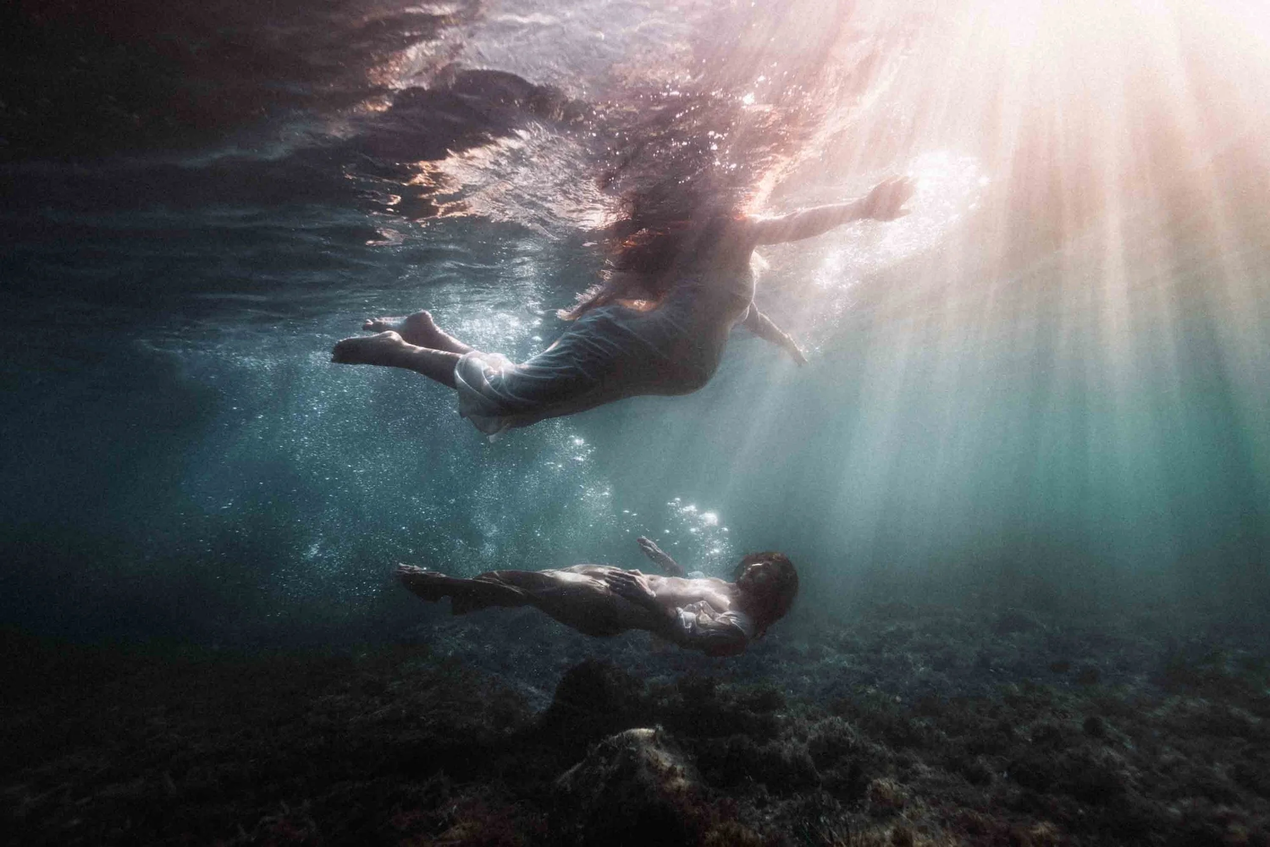 Deux personnes nagent sous l'eau, avec des rayons de lumière qui pénètrent dans l'eau depuis la surface.