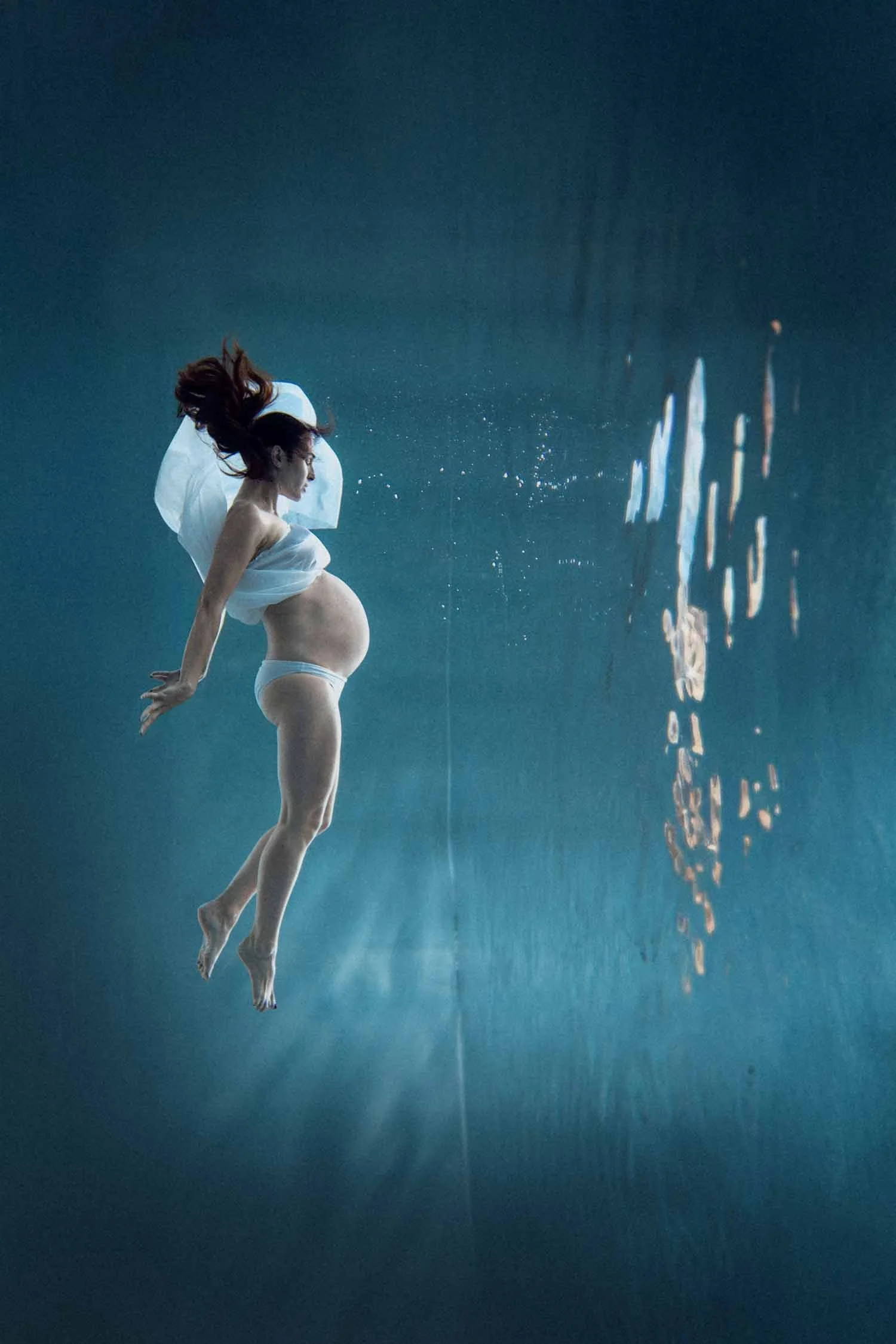 photo d'une femme enceinte sous l'eau dans une piscine bleue avec un voilage blanc, ventre dénudé