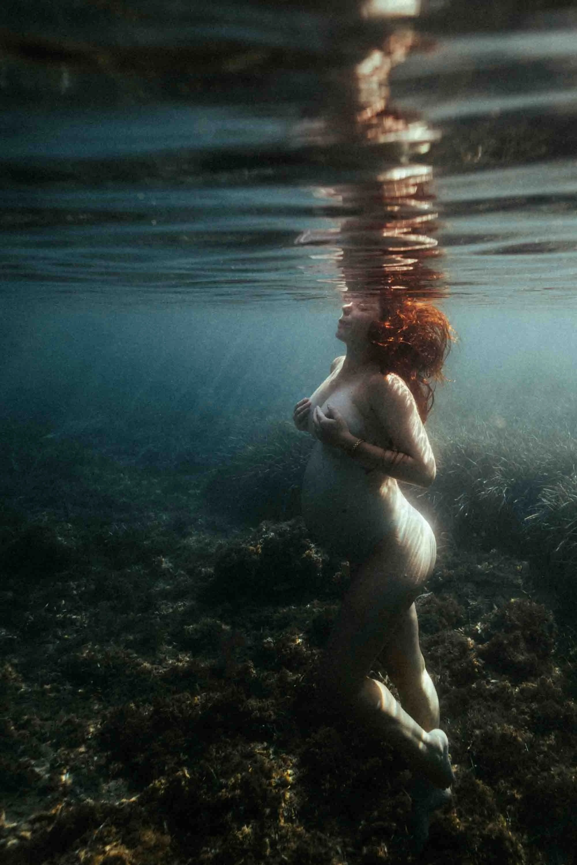Femme nue sous l'eau dans un milieu naturel avec des rochers et de la végétation.