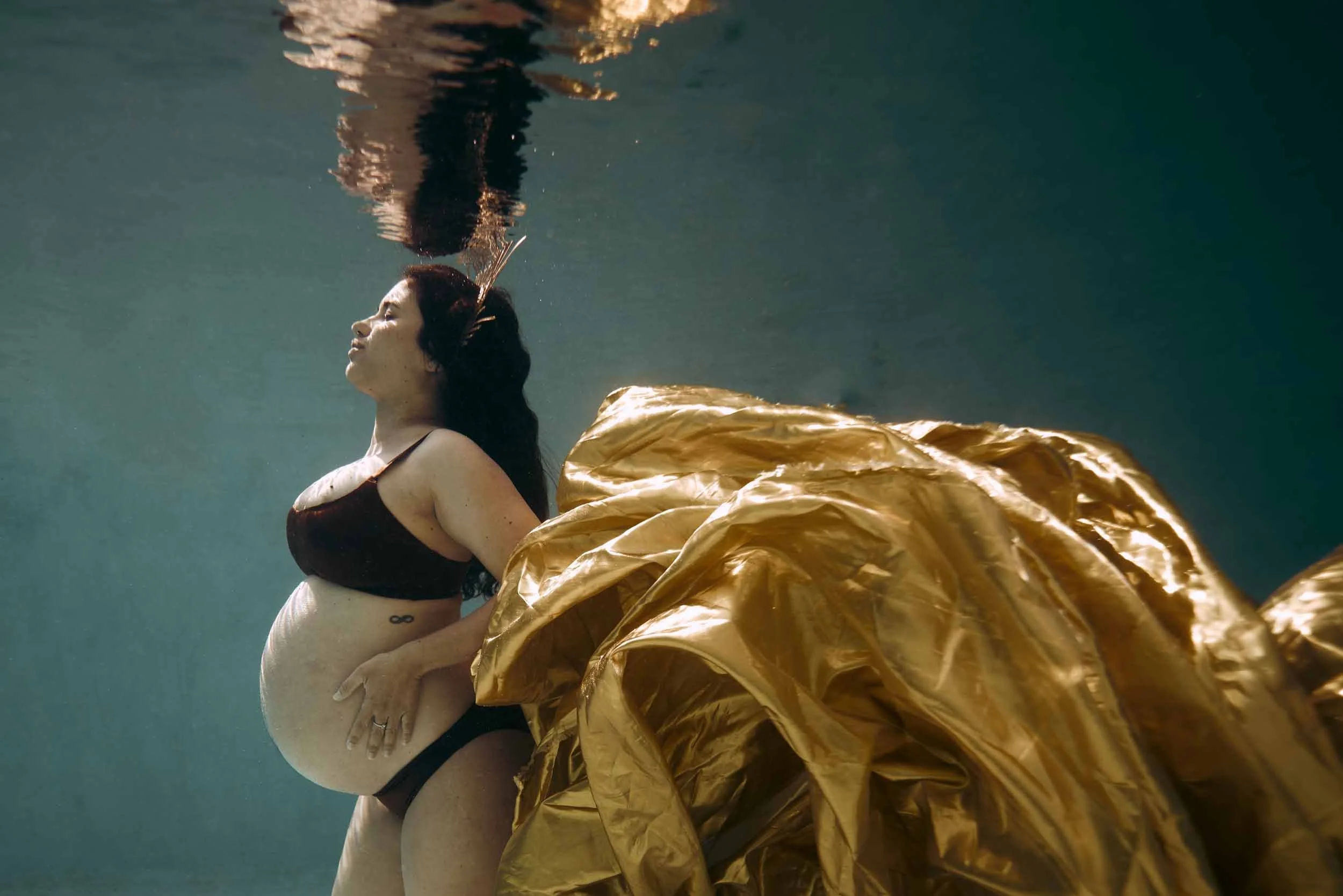 Une femme enceinte en lingerie noire, avec un voile noir dans les cheveux, posant sous l'eau avec un drapé doré à ses côtés.