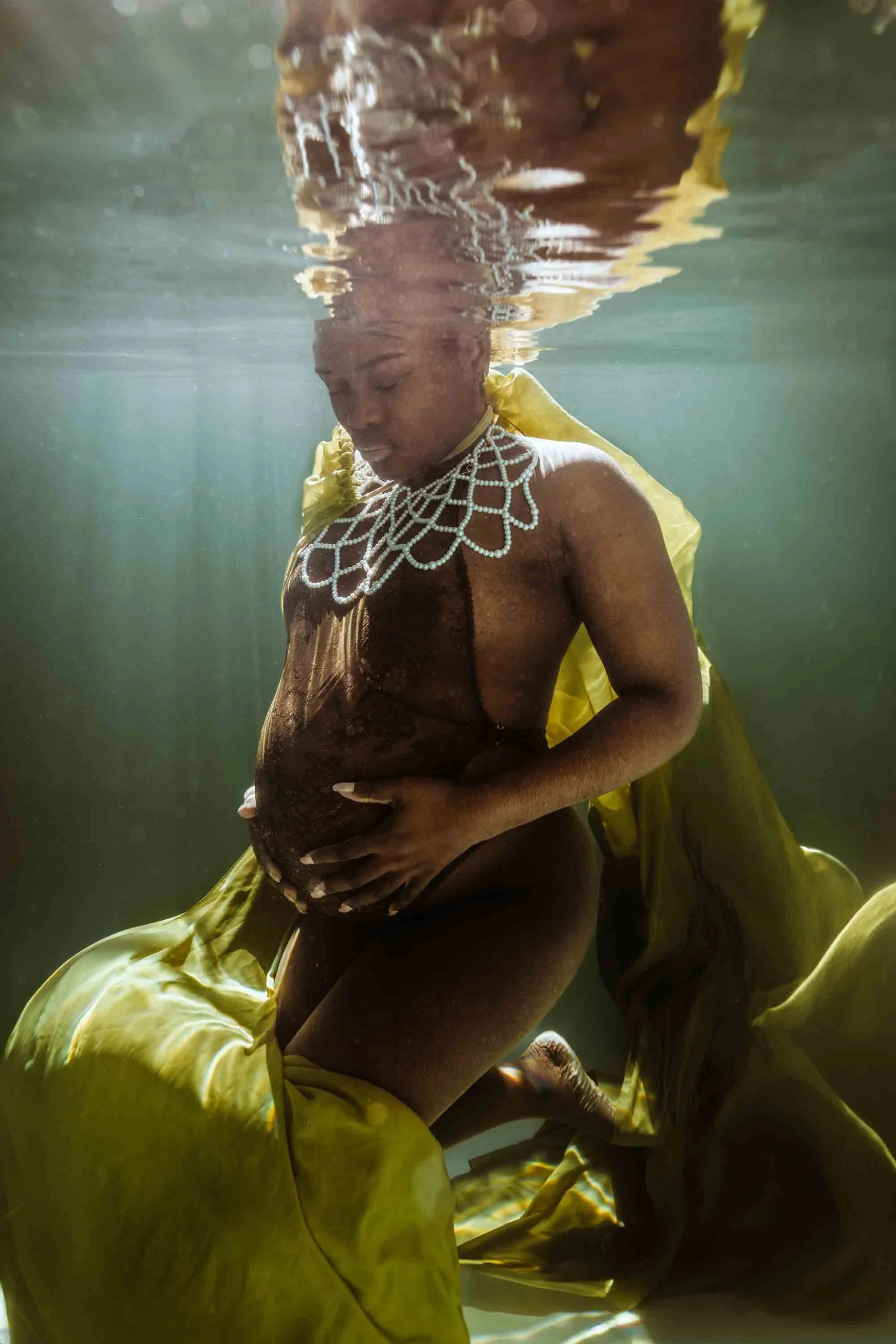Une femme enceinte en sous-vêtements, portant un collier en perles, posant sous l'eau avec une robe jaune, tenant son ventre rond et regardant vers le bas.