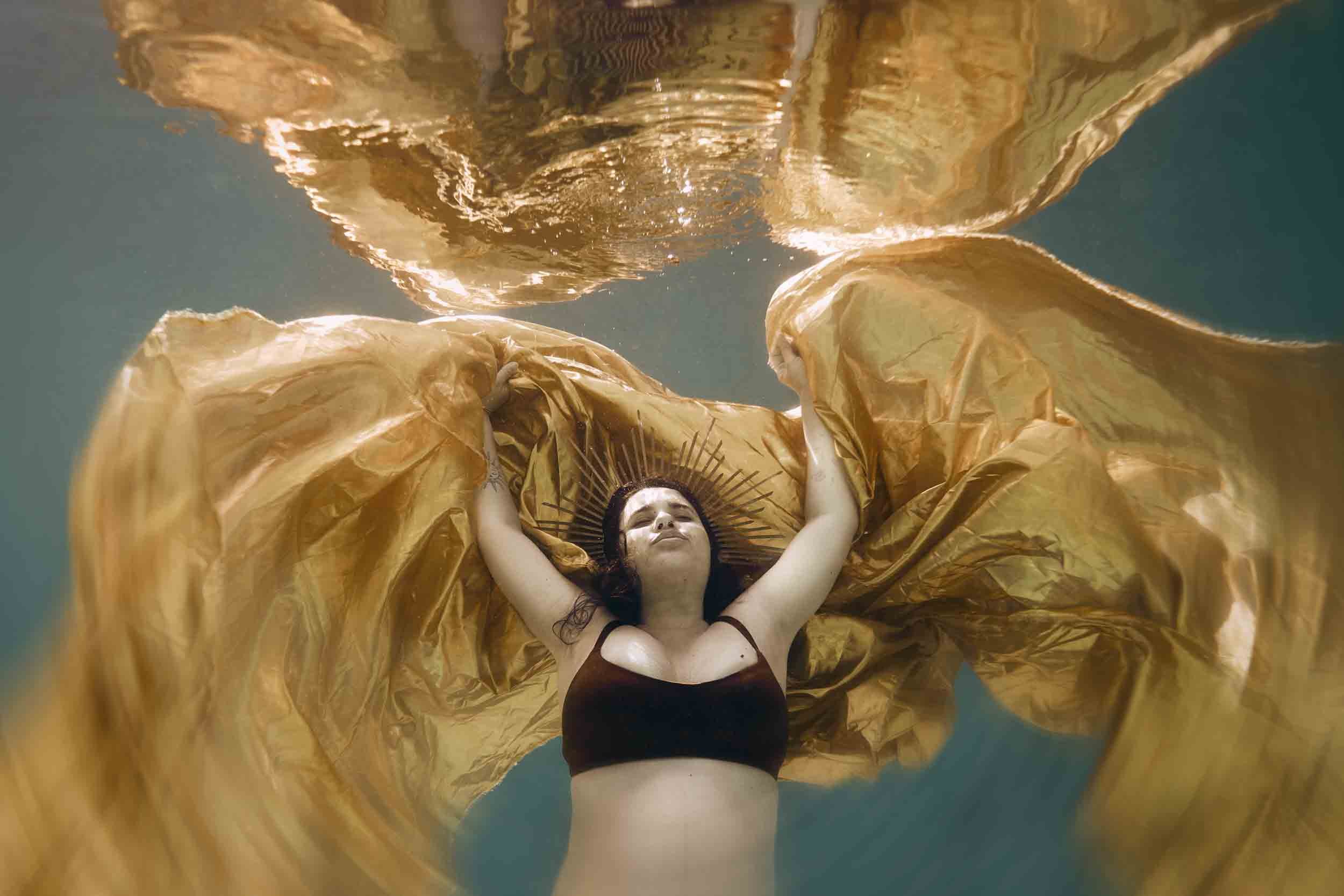 Une femme nage sous l'eau, portant une robe dorée et une couronne de style religieuse, avec des effets de lumière et de mouvement dans l'eau.