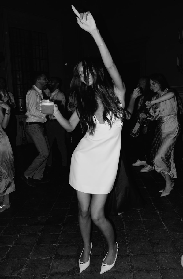 Black and white photo of people dancing at a party, featuring a woman in a white dress holding a drink and raising her arm.