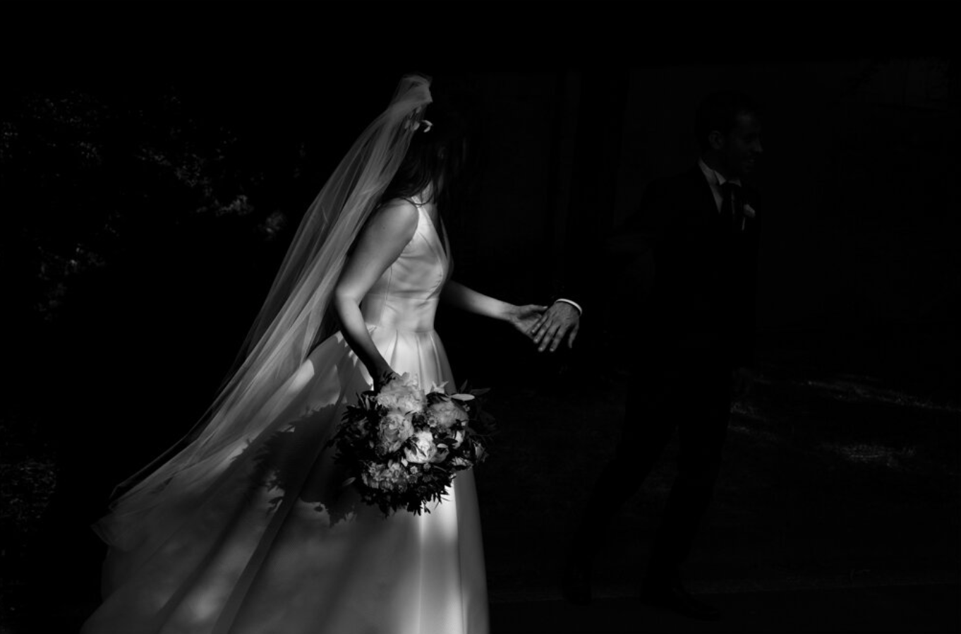 Black and white photo of a bride in a wedding dress holding a bouquet, walking with a groom.