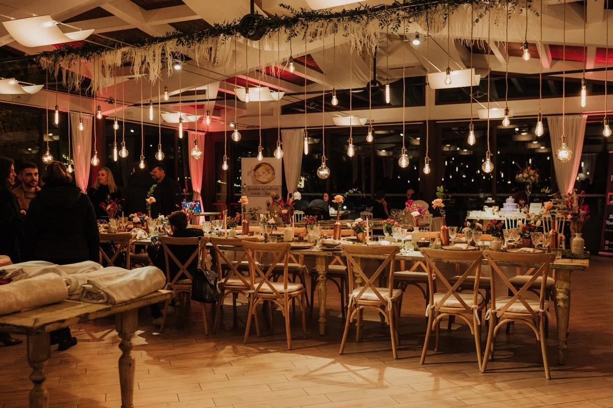 Decorative dining room with hanging light bulbs, wooden tables and chairs, floral centerpieces, and people socializing.