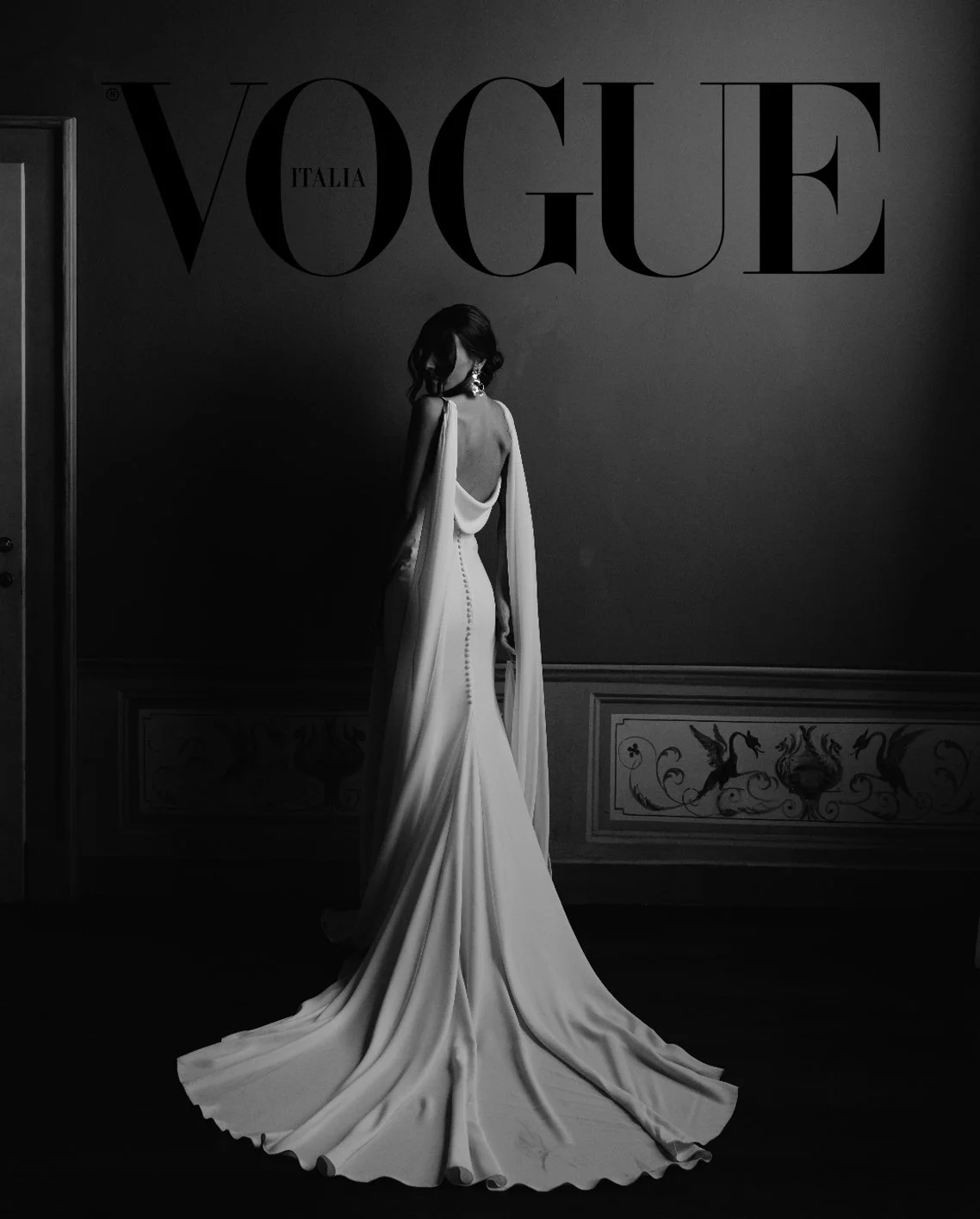 Black and white Vogue Italia cover featuring a woman in an elegant gown with a long train, standing in a dimly lit room and facing away.