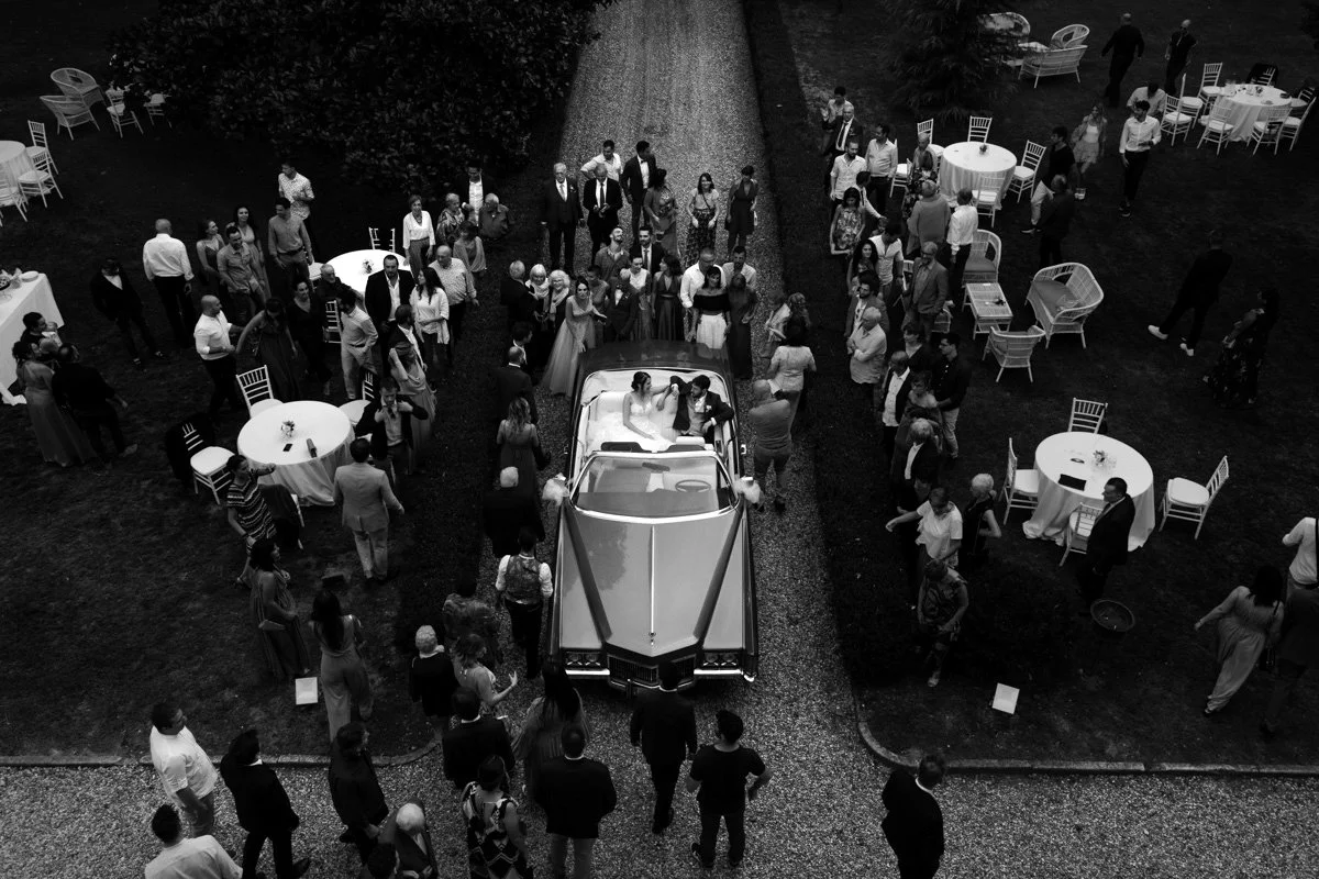 Black and white photo of a wedding scene with a couple in a vintage car surrounded by guests on a driveway.
