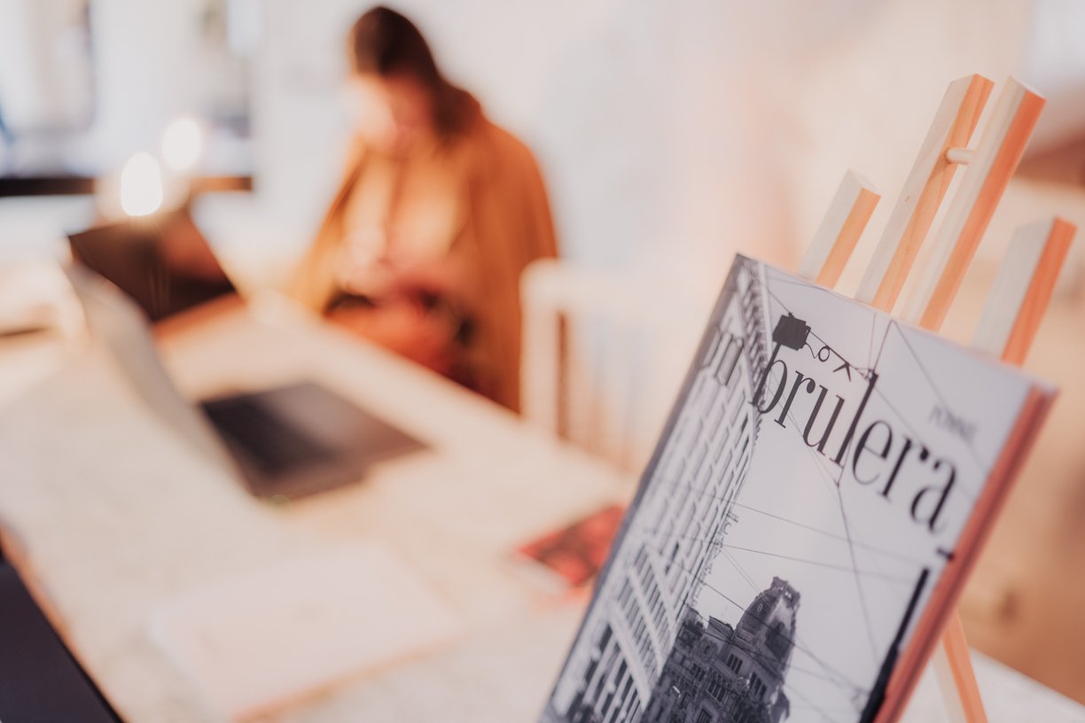 A desk with an open laptop, blurred background with a person using a smartphone, and a magazine on an easel in focus.