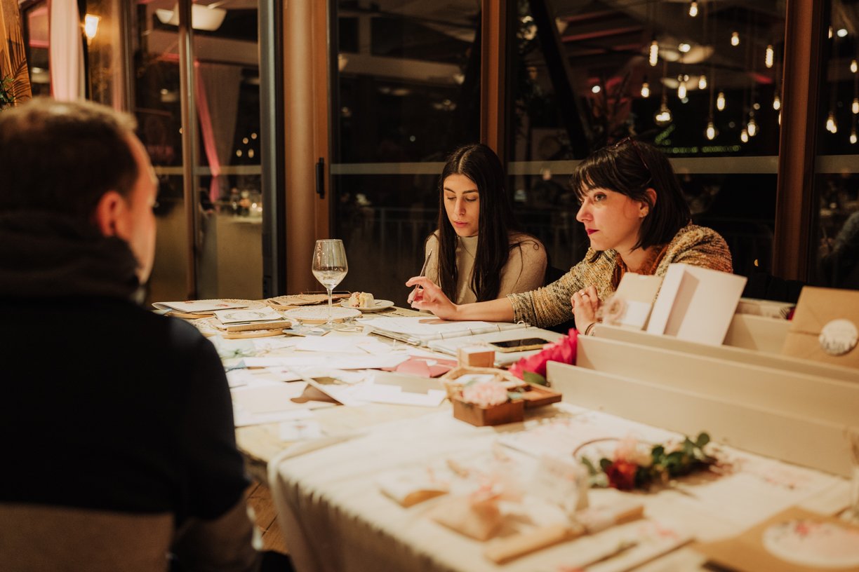 A group engaged in a creative meeting, featuring two women focused on planning with papers and documents spread across a table, as a man sits across from them in a warmly lit room.