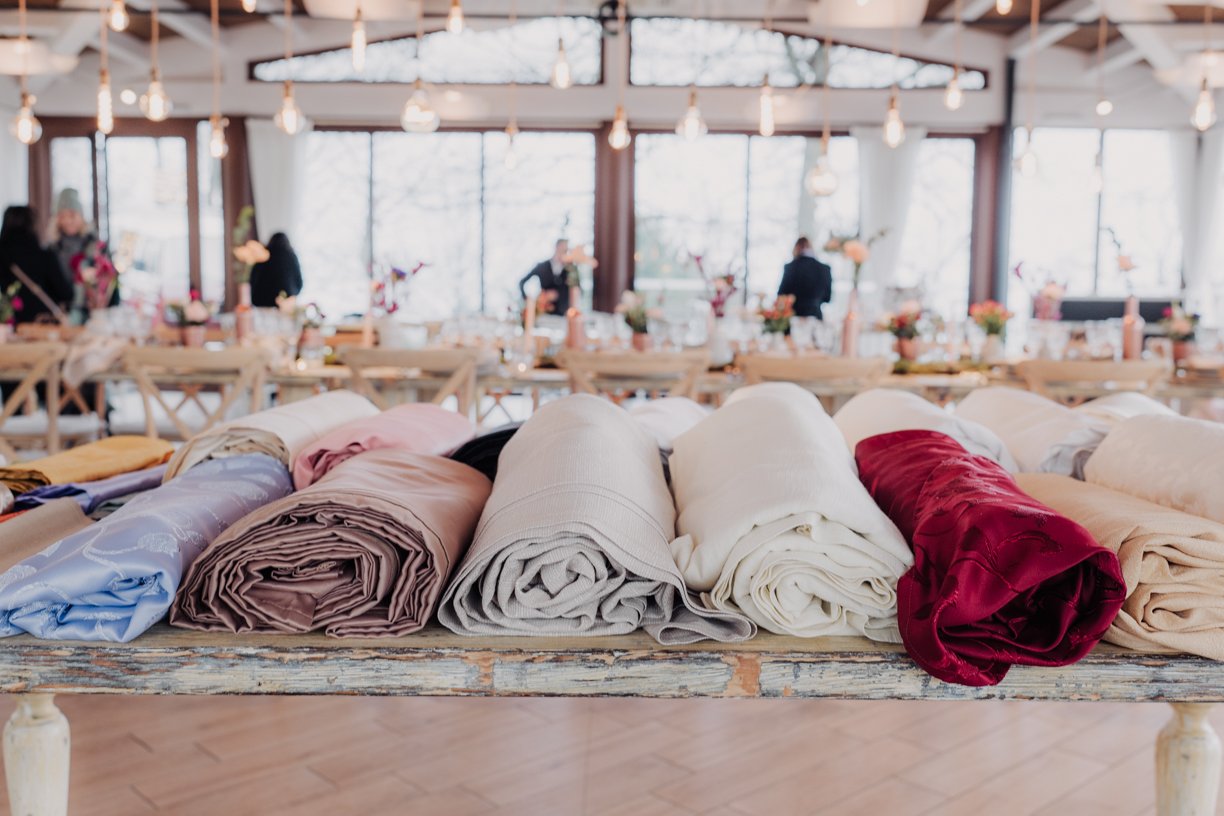 Rolled fabric bolts on a wooden table in a bright room with tables and pendant lights in the background.