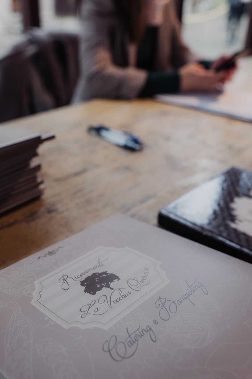 Close-up of a brochure on a wooden table with "Ristorante La Vecchia Quercia" and "Catering e Banqueting" written on it. A person is blurred in the background holding a smartphone.