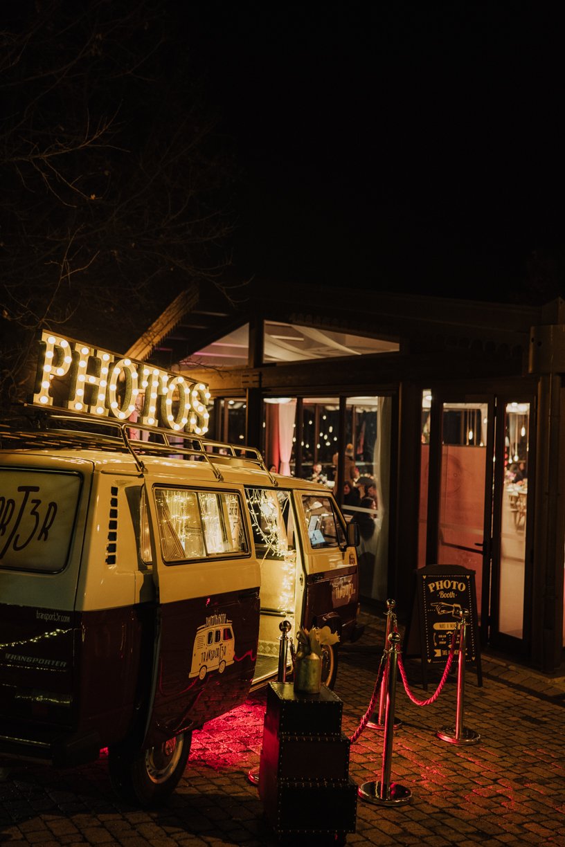 Retro van converted into a photo booth with illuminated 'PHOTOS' sign on top, parked outside a lit building at night.