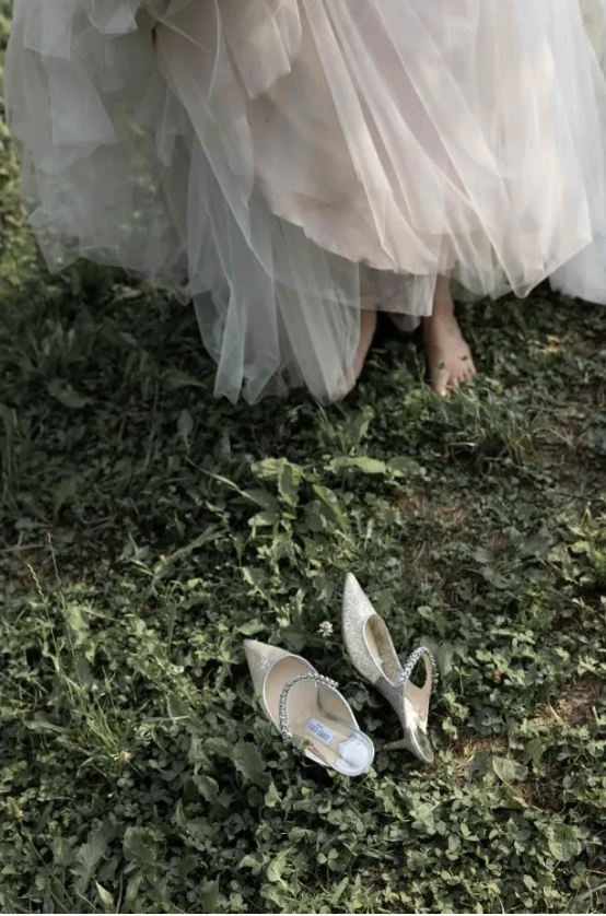 A person in a layered white dress stands barefoot on grass next to glittery high-heeled shoes.