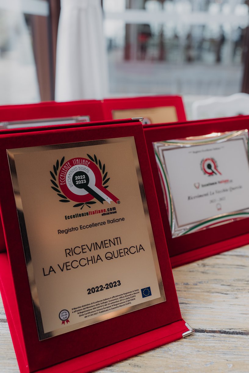 Awards in red frames displayed on a table, featuring text "Registro Eccellenze Italiane" and "Ricevimenti La Vecchia Quercia 2022-2023". The awards have a laurel wreath design with the year 2022-2023 and European Union logo.