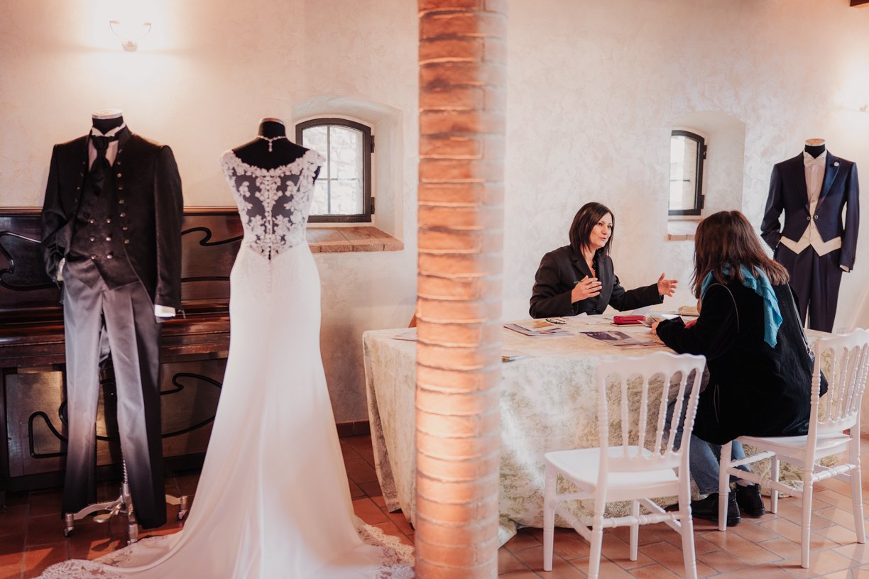 Two women sitting at a table discussing in a room with displayed wedding attire, including a bridal gown and suits on mannequins.