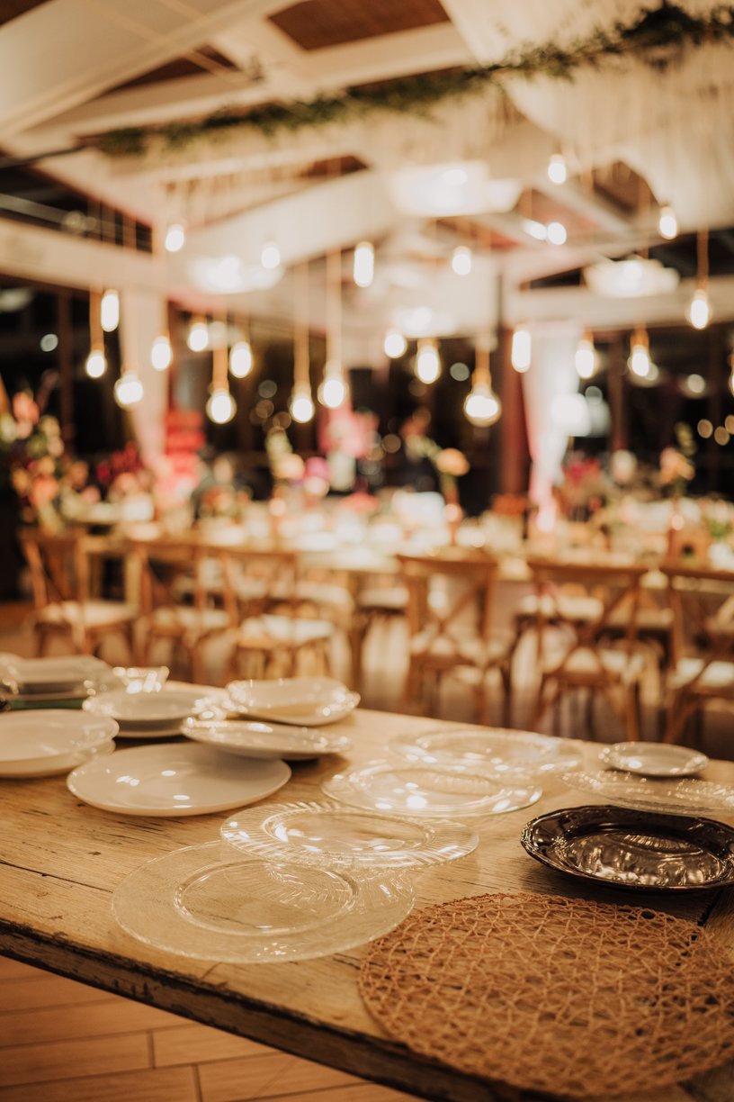Elegantly set dining table with plates and woven placemat, blurred background with hanging lights and floral decor.
