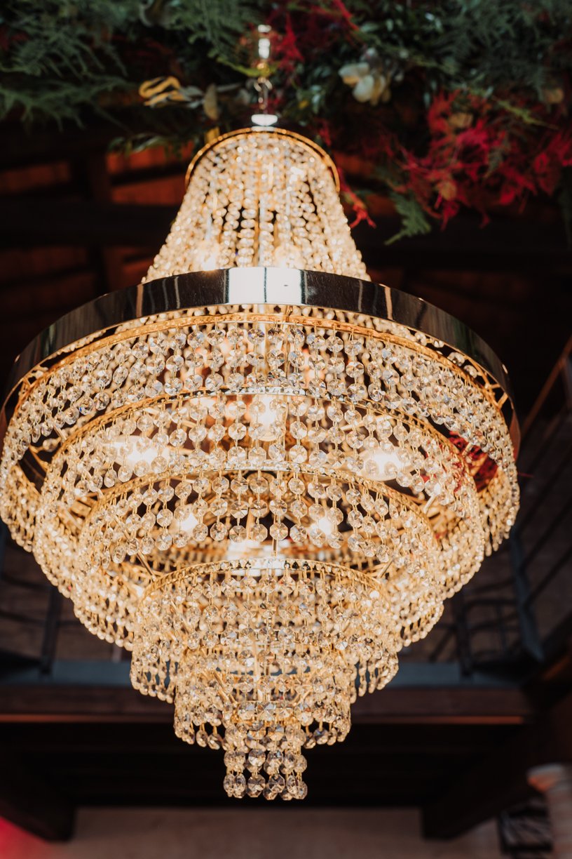 Elegant crystal chandelier hanging from a decorated ceiling, featuring numerous sparkling crystals and golden fixtures, with floral decorations above.