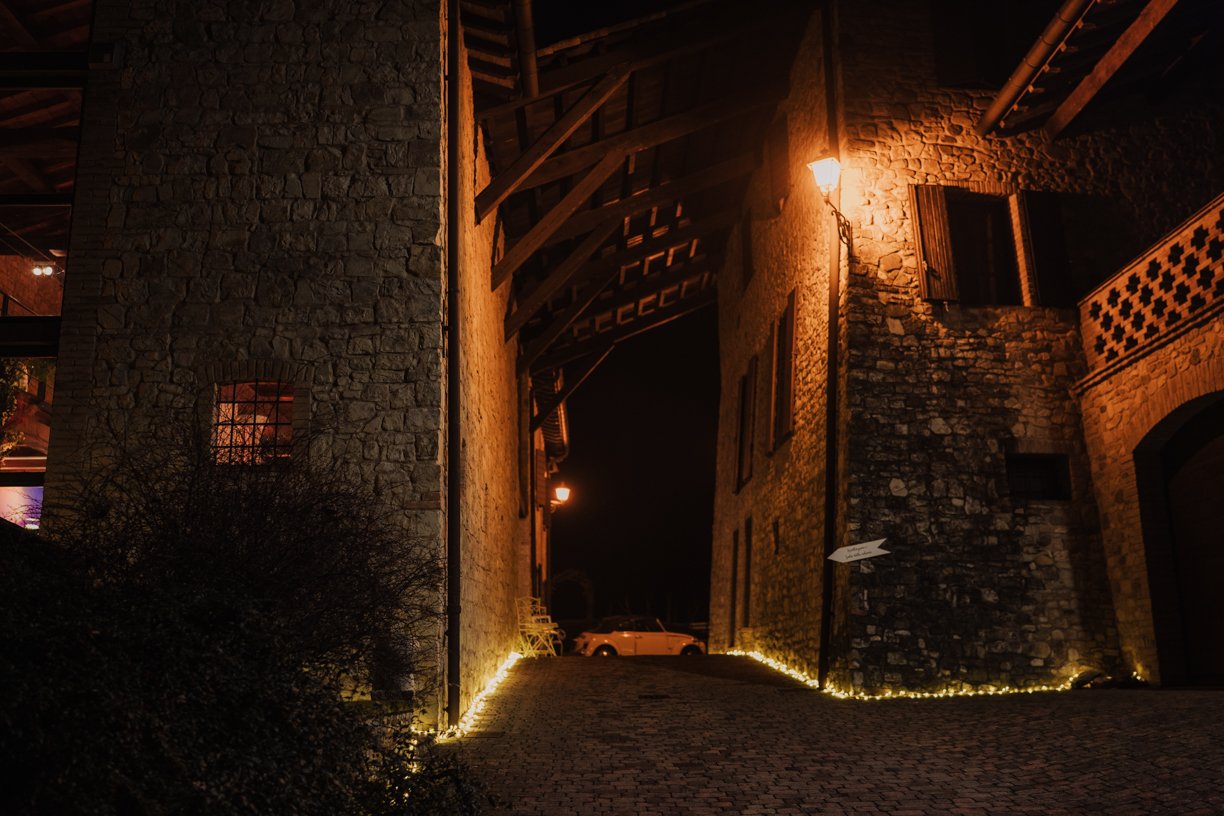 Stone building with warm lighting at night, cobblestone pathway, and a parked car in the background.